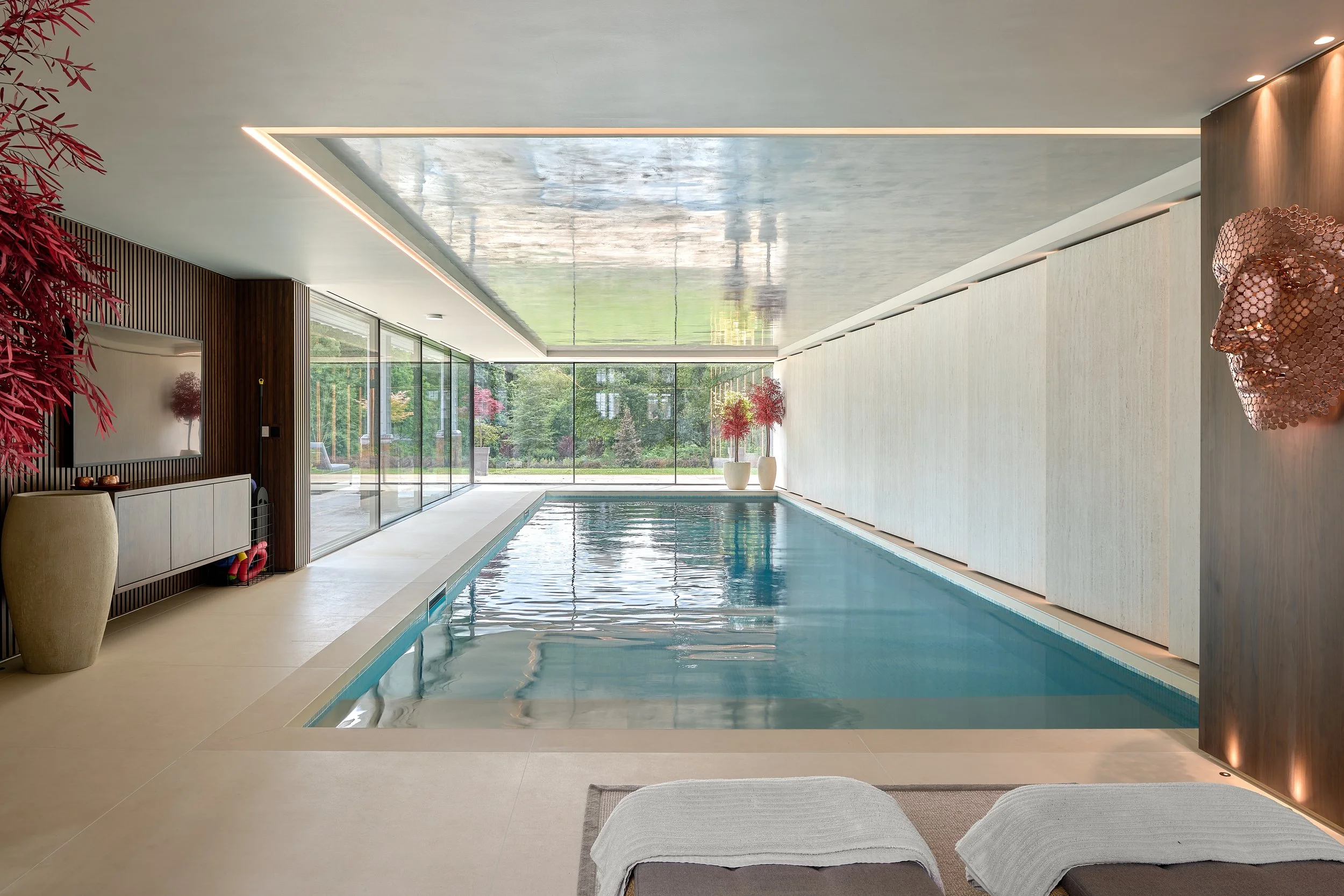 Indoor swimming pool with large glass windows showing greenery outside, modern decor, and a pink textured face sculpture on the wall.