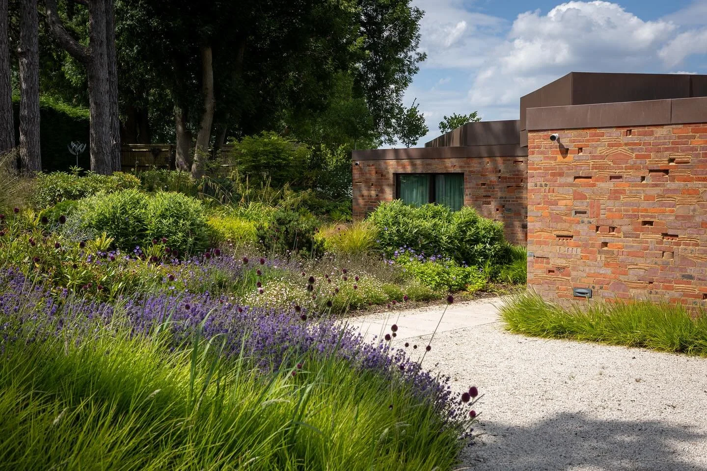 A brick house with a gravel pathway surrounded by lush green plants and purple flowers, with tall trees in the background and a partly cloudy sky above.