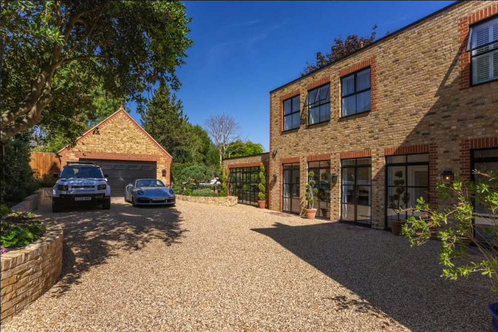 A gravel driveway in front of a brick house with large windows, potted plants, and a garage with two cars parked outside under a tree on a sunny day.