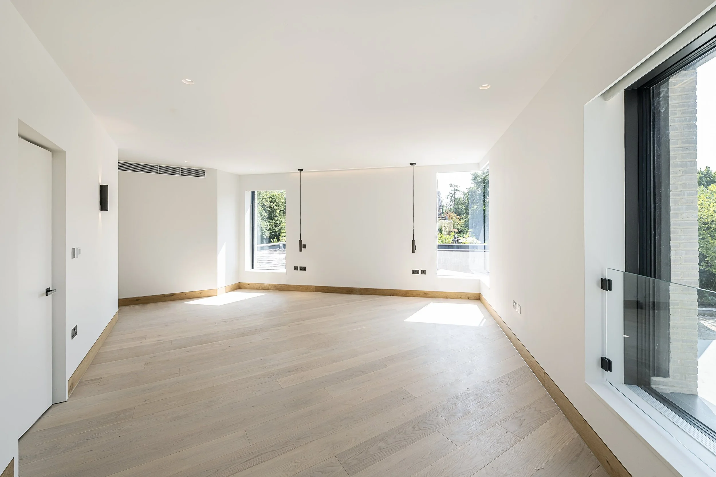 Empty modern living room with white walls, light wood flooring, large windows letting in natural light, and minimalistic pendant lighting fixtures.