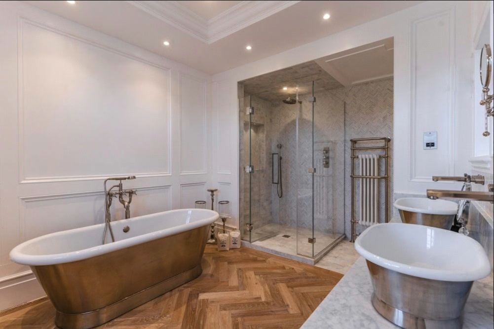 Luxury bathroom featuring a freestanding copper bathtub, a glass-enclosed shower with rainfall and handheld showerheads, a marble countertop with two vessel sinks, wooden herringbone floor, and elegant white wall paneling.