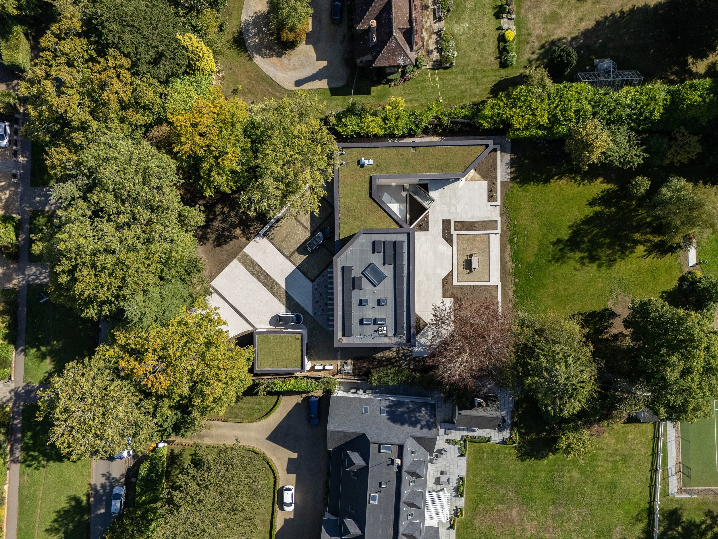 An aerial view of a residential house with a flat roof, surrounded by trees and a lawn, with cars parked in the driveway.