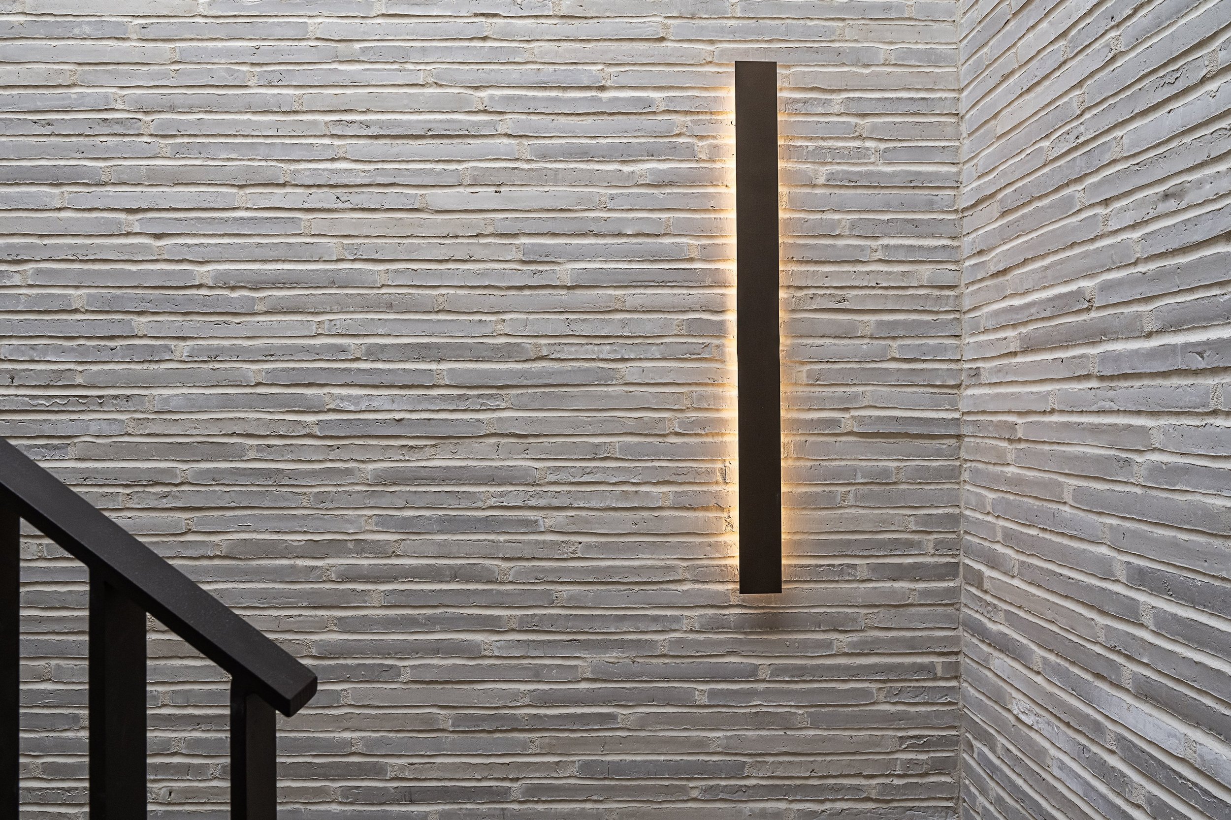 Modern black wall sconce with warm backlighting mounted on a textured white brick wall in a stairwell.