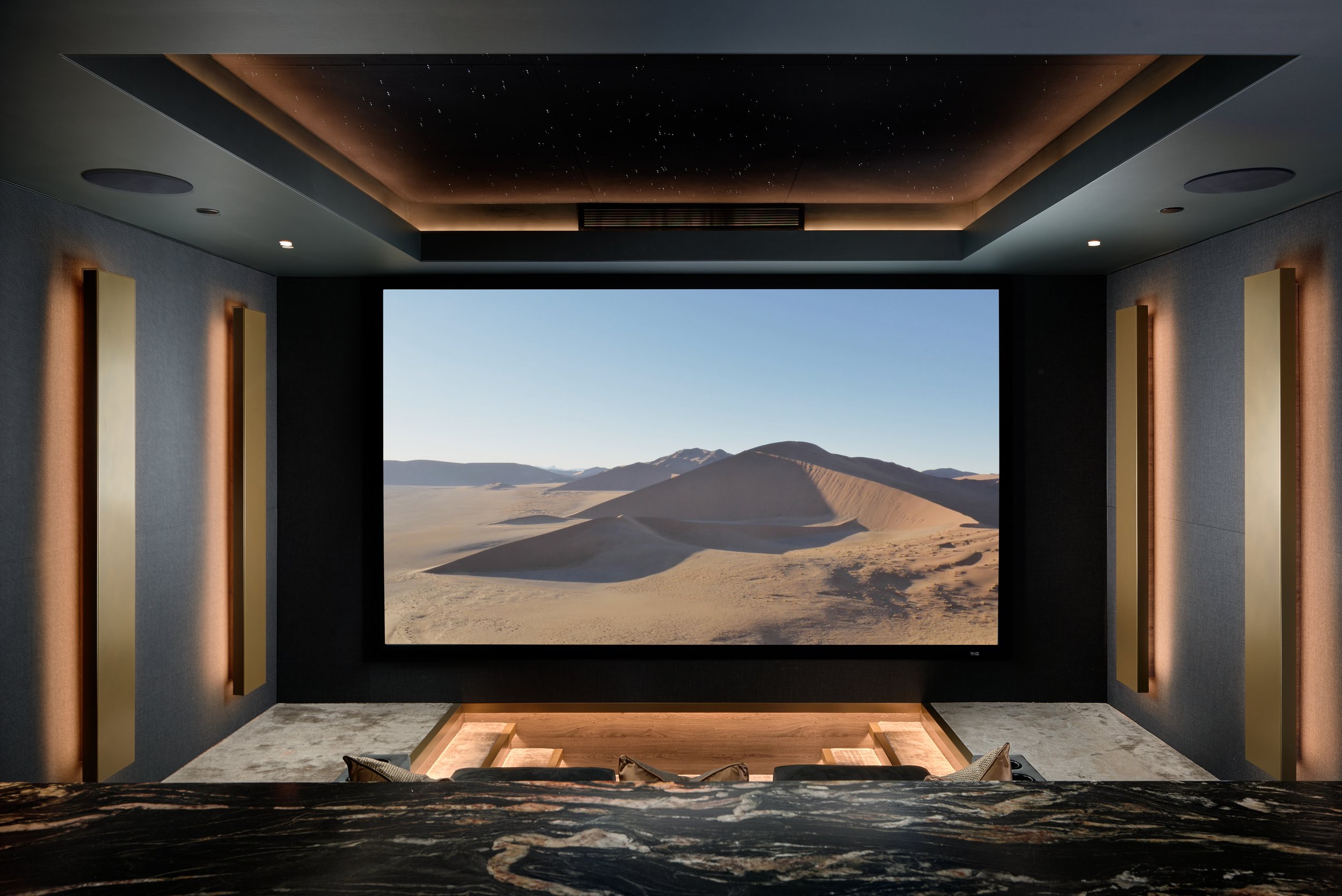 Modern home theater room with a large screen showing desert landscape with sand dunes and mountains, dark walls, and ambient lighting.