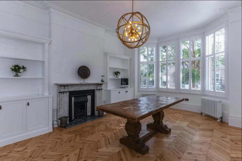 Living room with white walls, a fireplace with a marble mantel, built-in shelves, a large wooden table, a modern chandelier, and a bay window with white shutters.