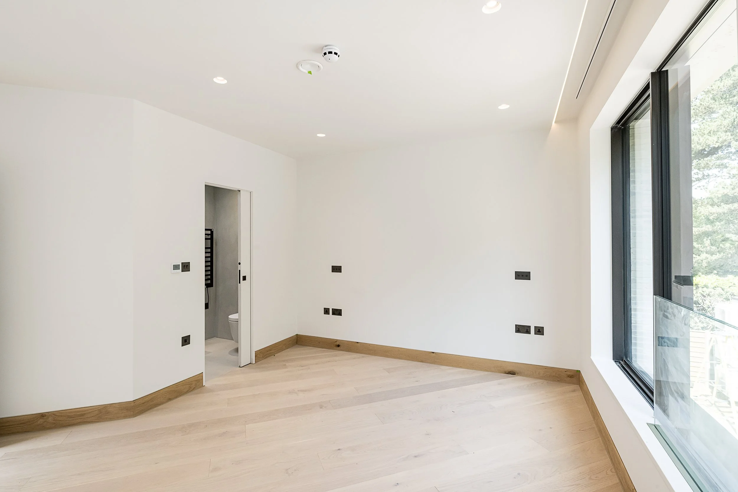 Empty room with white walls, light wood flooring, large window, and a small door leading to a bathroom.