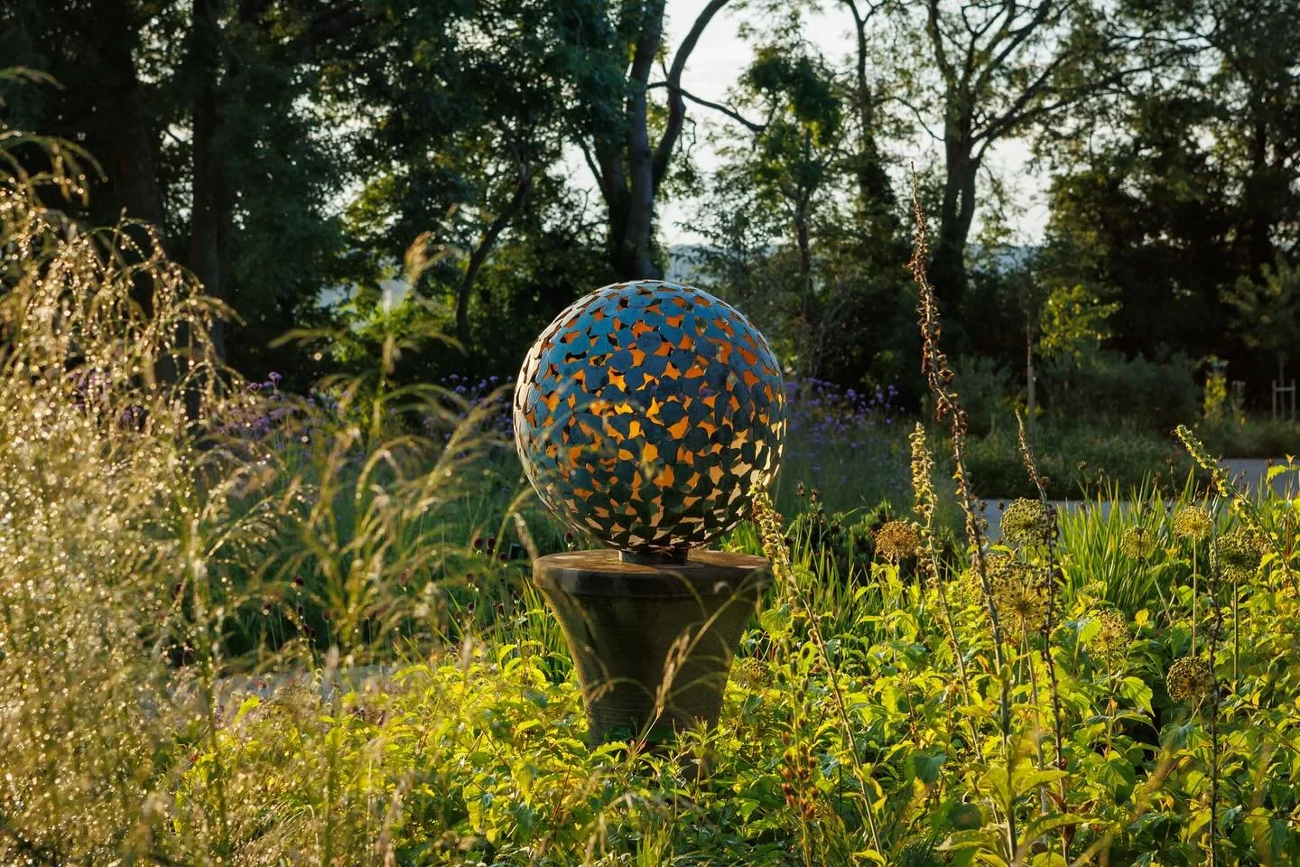 A decorative spherical sculpture with colorful scales on a pedestal in a lush garden at sunset.