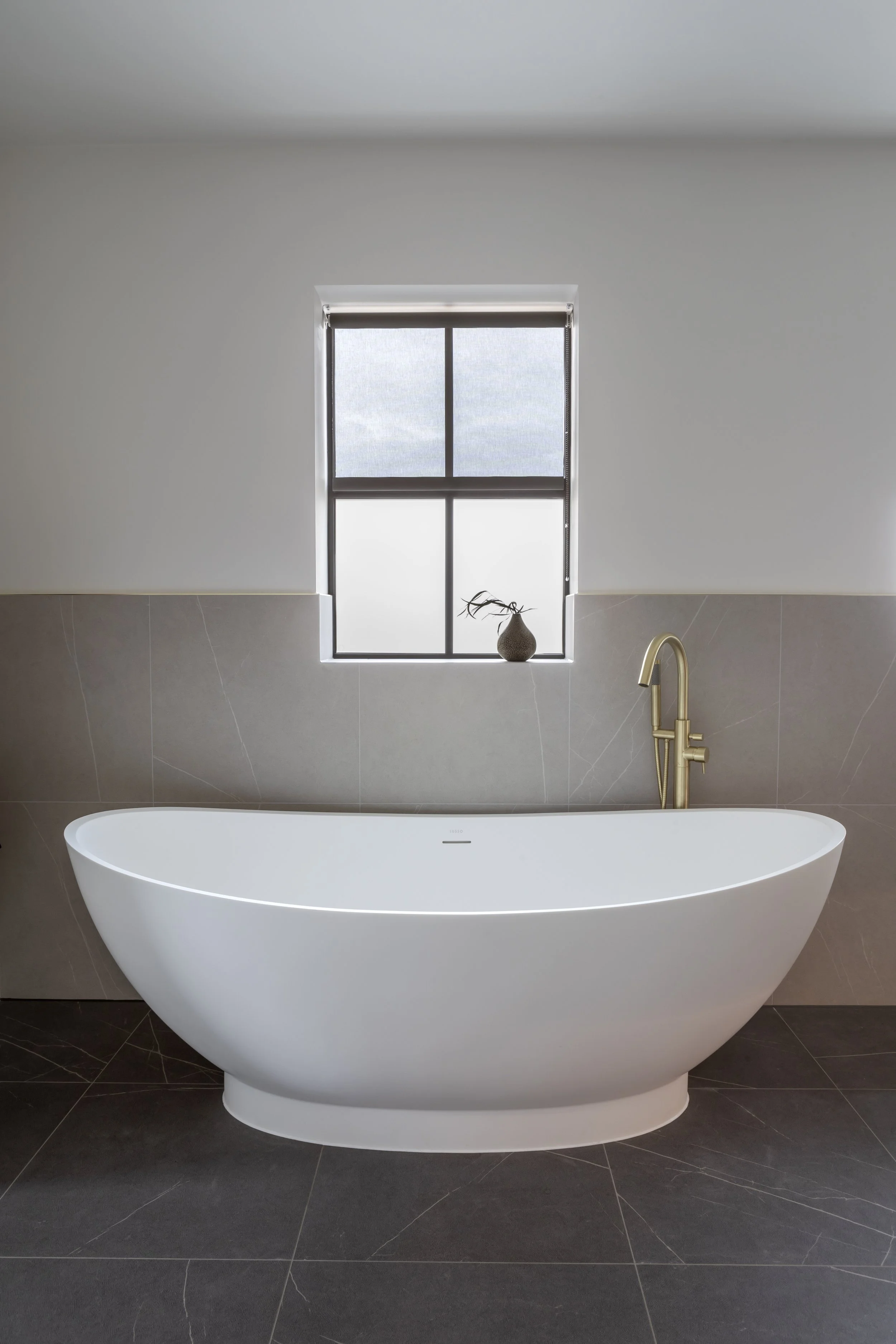 Modern bathroom with freestanding white bathtub, gold faucet, and small window with a decorative vase and plant on the windowsill.