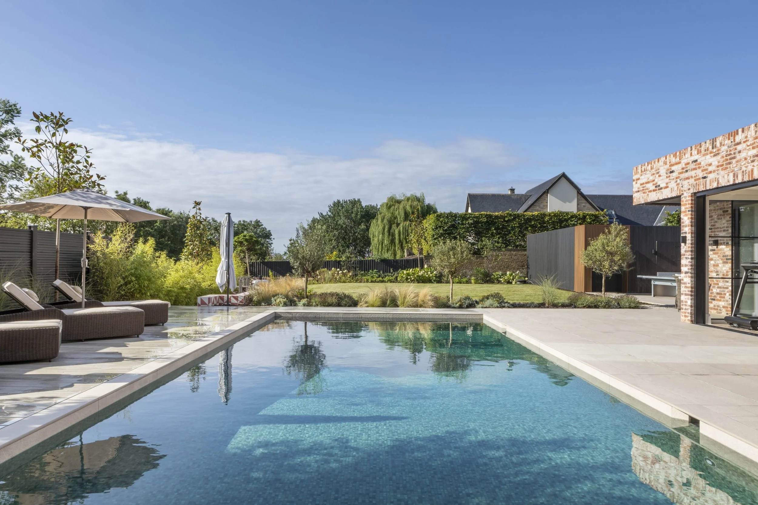 A backyard with a swimming pool, lounge chairs with umbrellas, a grassy area with trees, and a modern brick house on a sunny day.