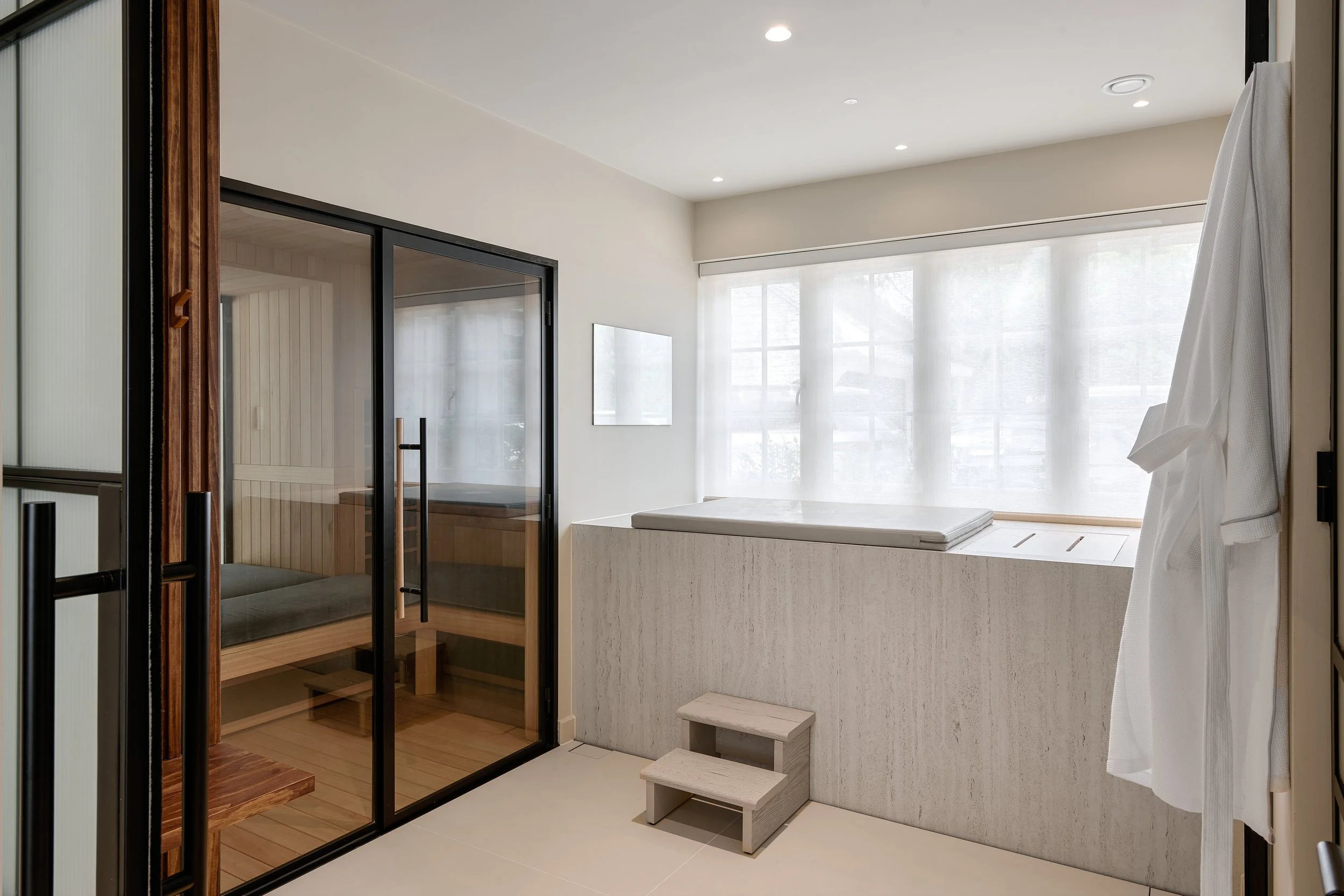 Modern bathroom with a large soaking tub, small wooden steps, a glass-enclosed dry sauna, and natural light coming through sheer curtains.