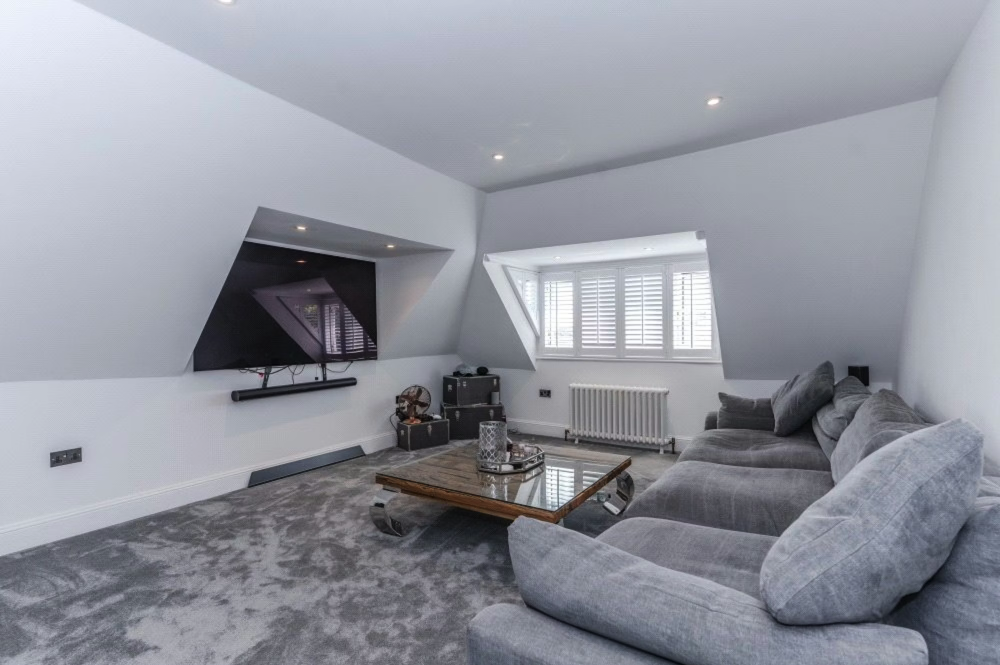 Living room with a gray sofa, a flat-screen TV mounted on the wall, and a wooden coffee table on a gray carpet, with windows and shutters on the wall.