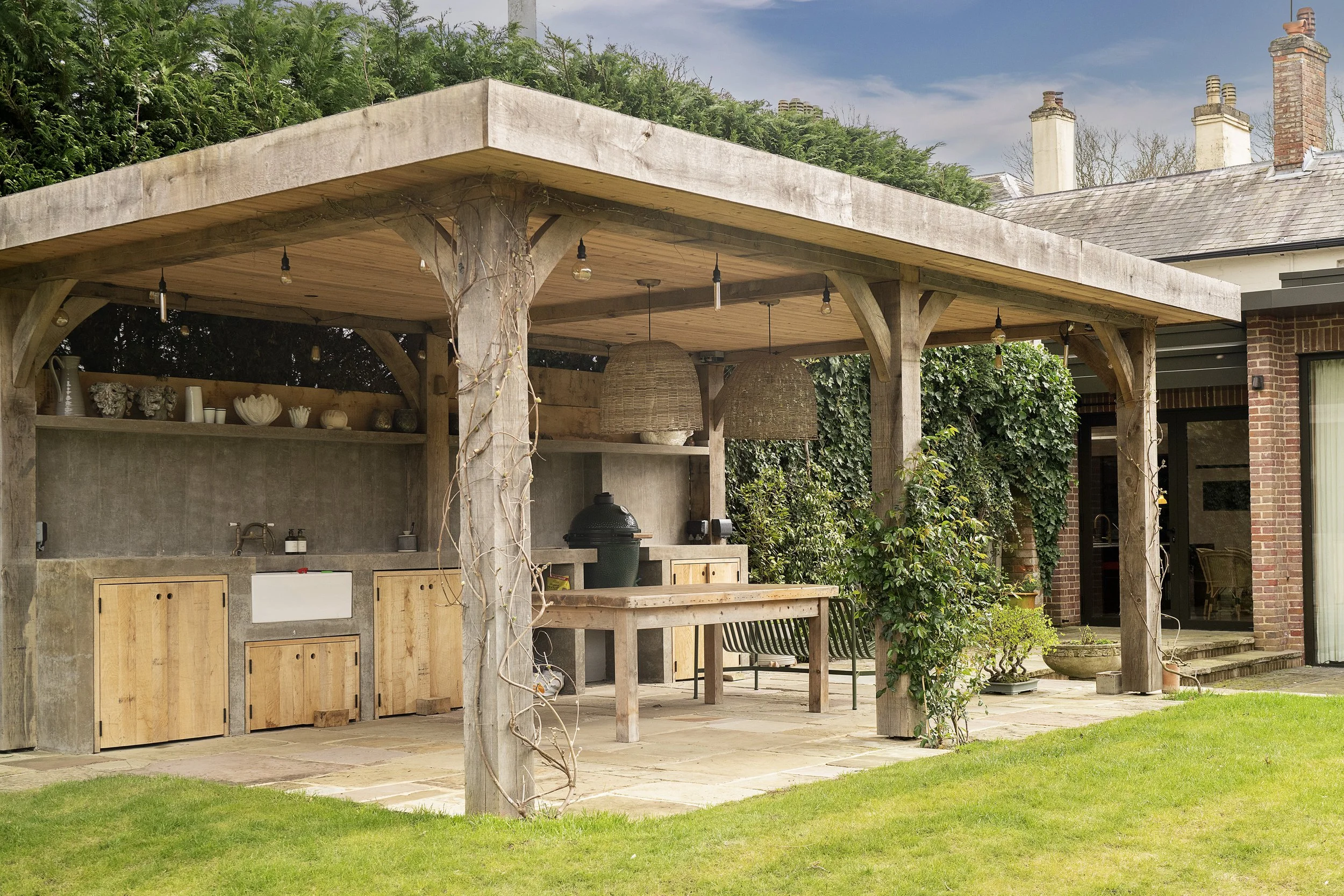 Outdoor kitchen with wooden pergola, built-in sink, countertop, cabinets, hanging woven light fixtures, and a green ceramic grill, set on a stone patio with a lush green lawn.