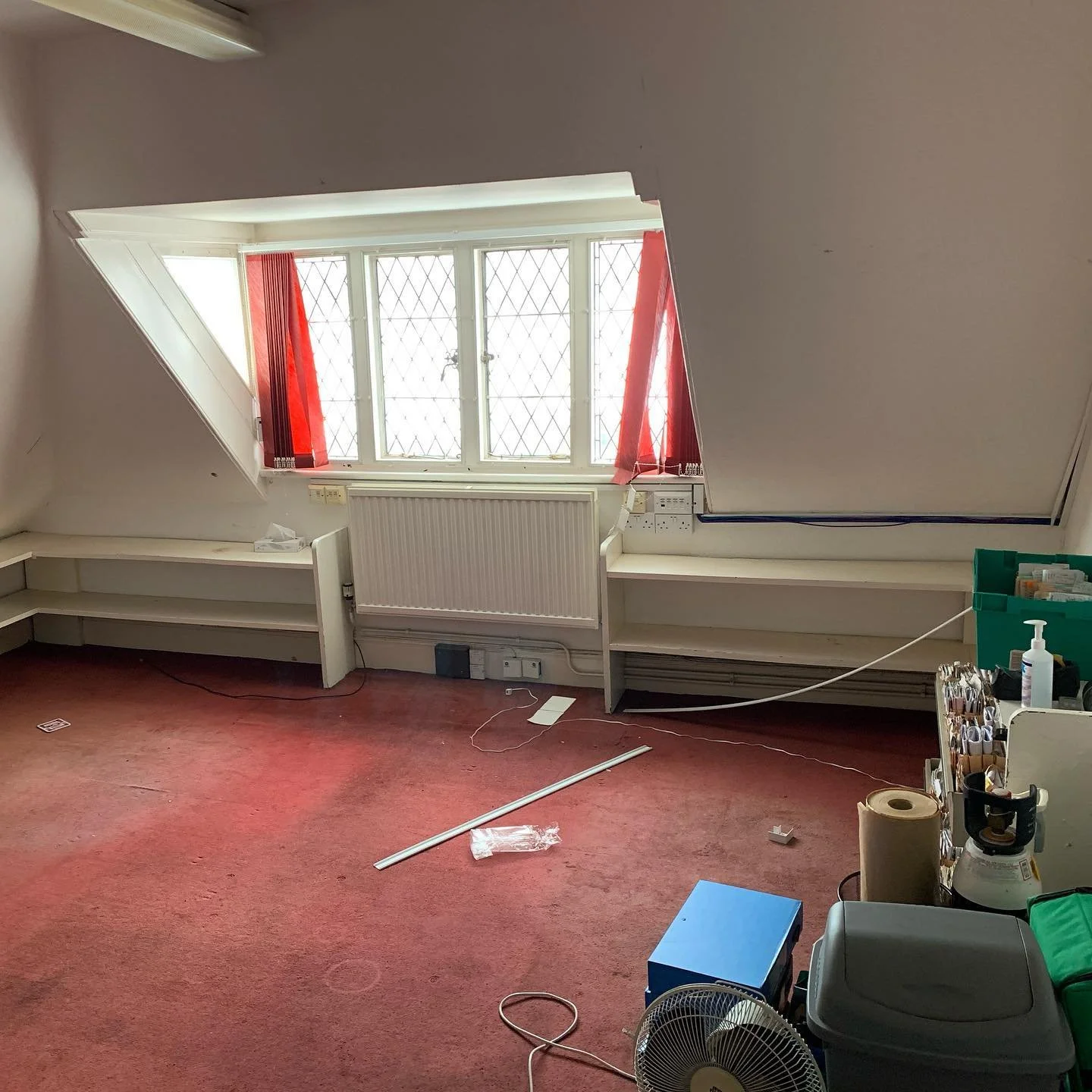 An empty room with a sloped ceiling and a large window with red curtains, a radiator, and various items on the floor including a fan, a blue box, a roll of paper towels, and some cables.
