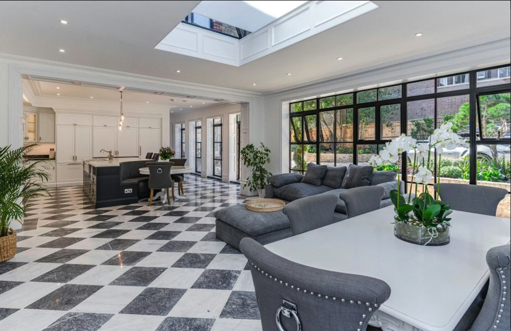 Spacious open-plan living and dining area with black and white checkered marble flooring, gray sofas, large windows, and a bright kitchen with an island.
