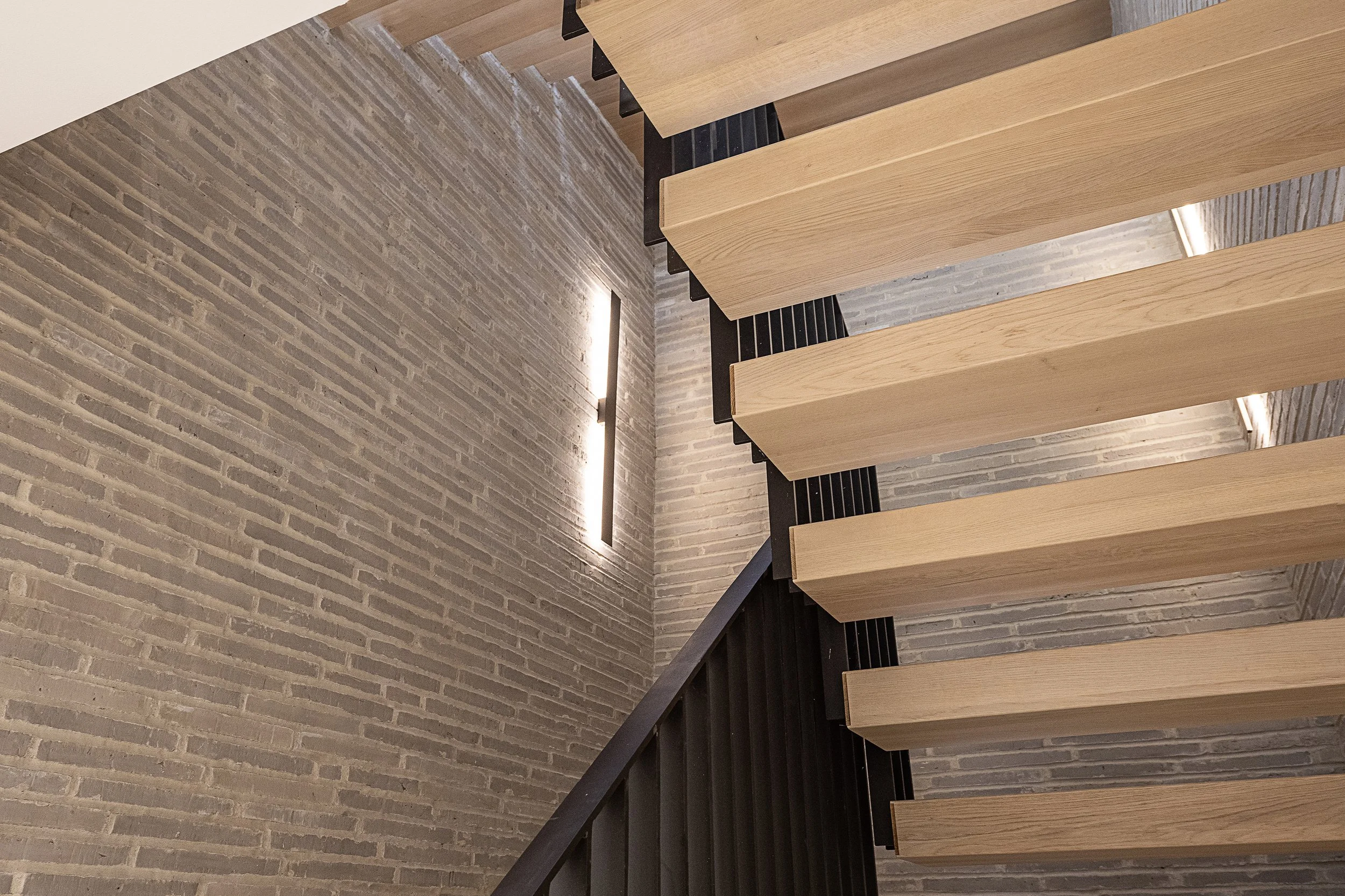 Interior view of a modern staircase with hanging wooden steps, black metal supports, and a brick wall with wall-mounted lighting.