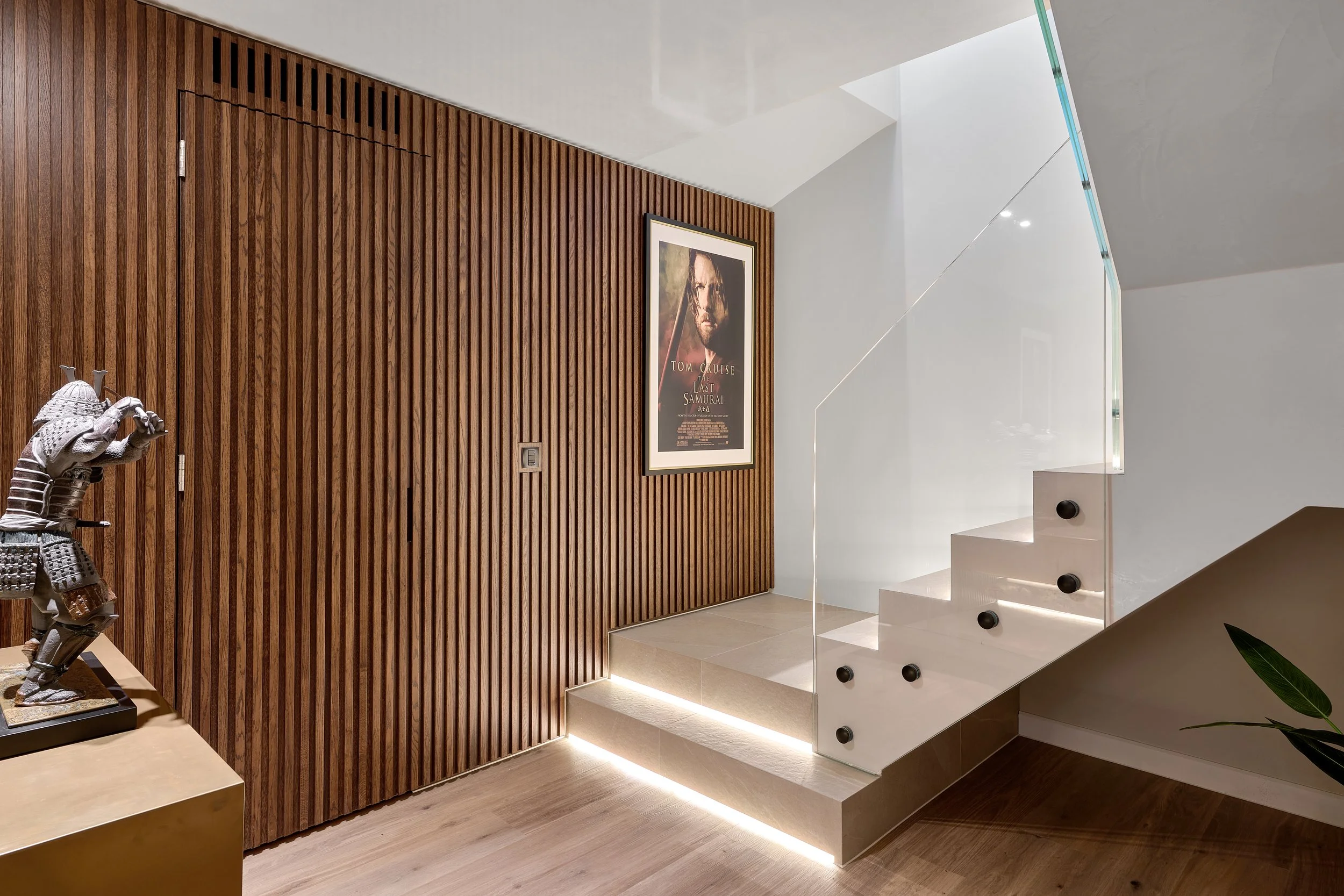 Modern interior staircase with white steps, black knobs, a glass railing, and LED lighting, adjacent to a wooden-paneled wall with a framed poster for the movie "Last Samurai" and a sculpture of a samurai in armor on a table.