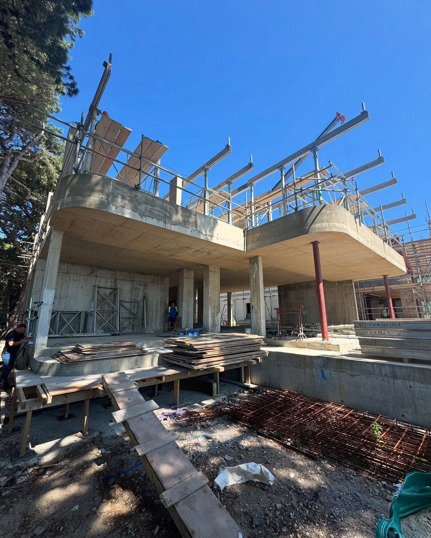 Construction site of a building with concrete structure, scaffolding, and construction materials, under a clear blue sky.