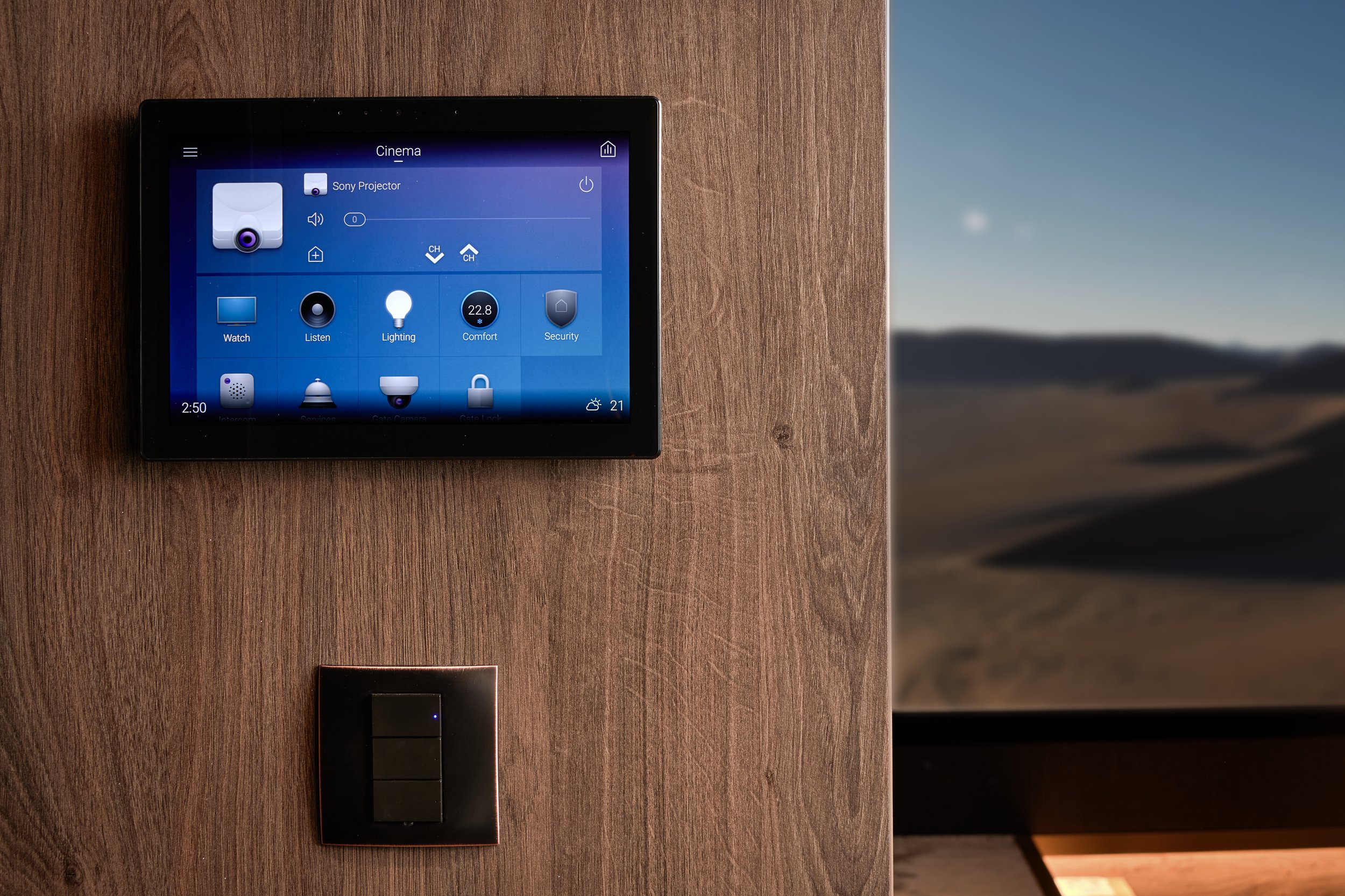 A wall-mounted smart home control panel displaying climate settings and a switch below it, with a window or screen showing an outdoor landscape with hills and a clear sky.