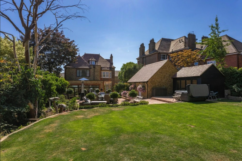 Backyard garden with green lawn, trees, shrubs, outdoor seating area with chairs and table, patio, and several houses with brick walls and chimneys under a clear blue sky.