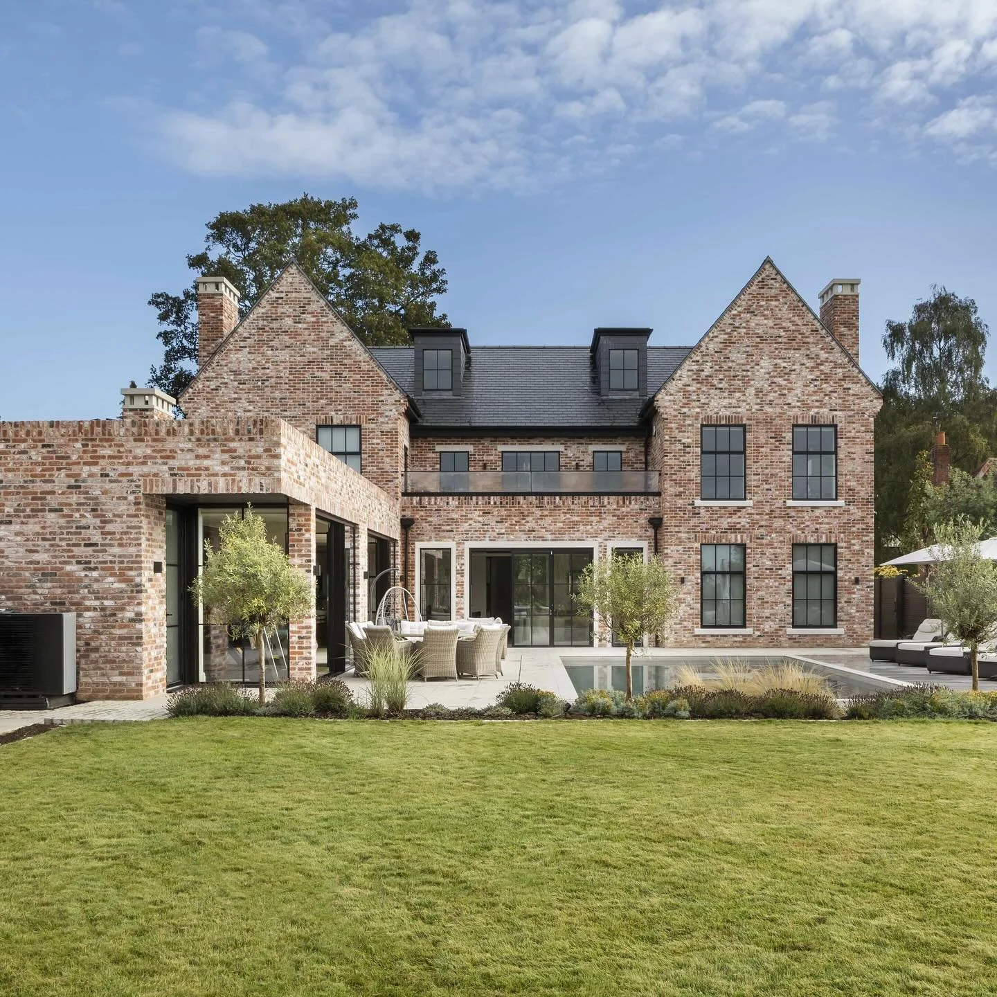 Back of a brick house with large windows, a patio area with outdoor furniture, and a swimming pool, surrounded by a well-maintained lawn and trees.