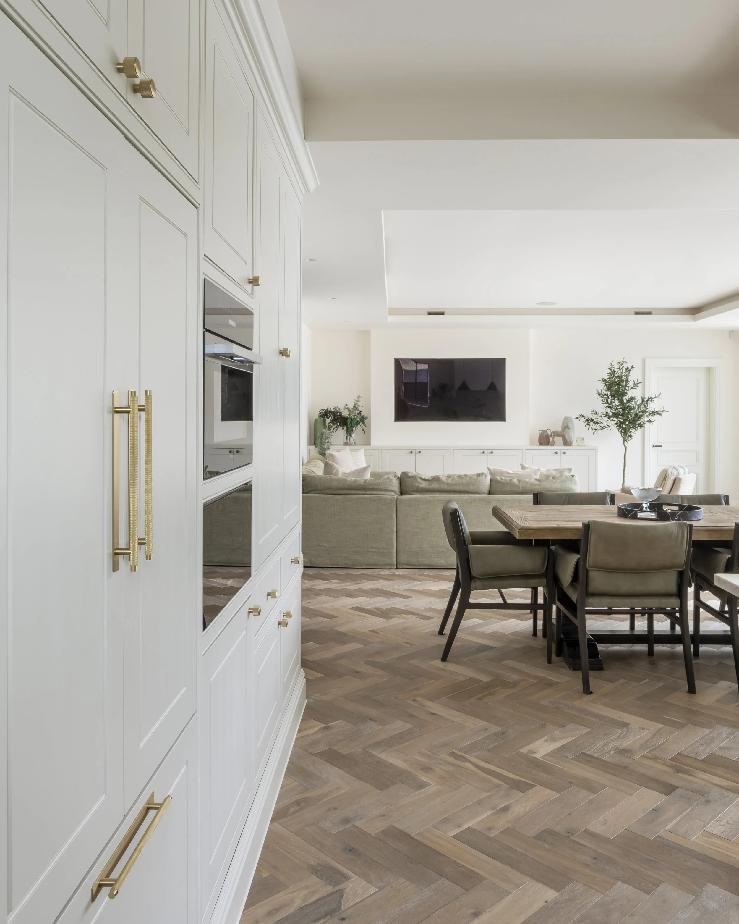 Open-plan living and dining room with a white built-in cabinetry on the left, a beige sofa facing a wall-mounted flat-screen TV, a wooden dining table with six chairs, and decor including plants and vases. The floor has a herringbone pattern with woo