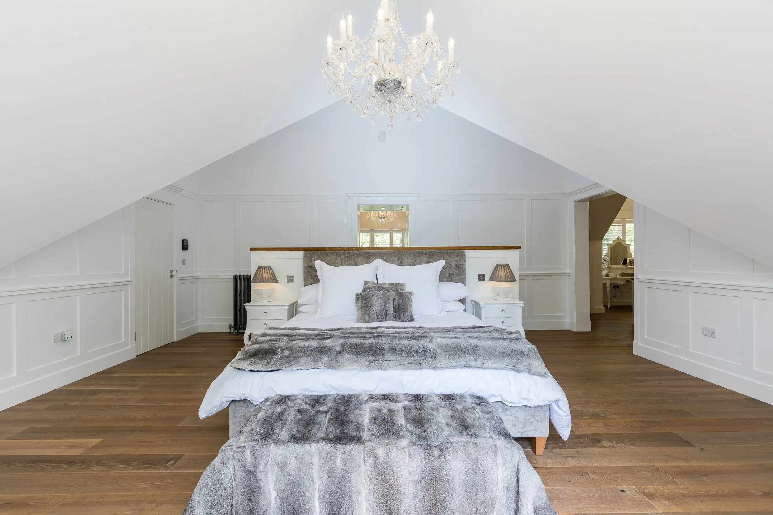 A bedroom with white walls, hardwood floors, a gray upholstered headboard, and a bed with white and gray bedding. There are two small white nightstands with lamps, and a chandelier hanging from the ceiling.