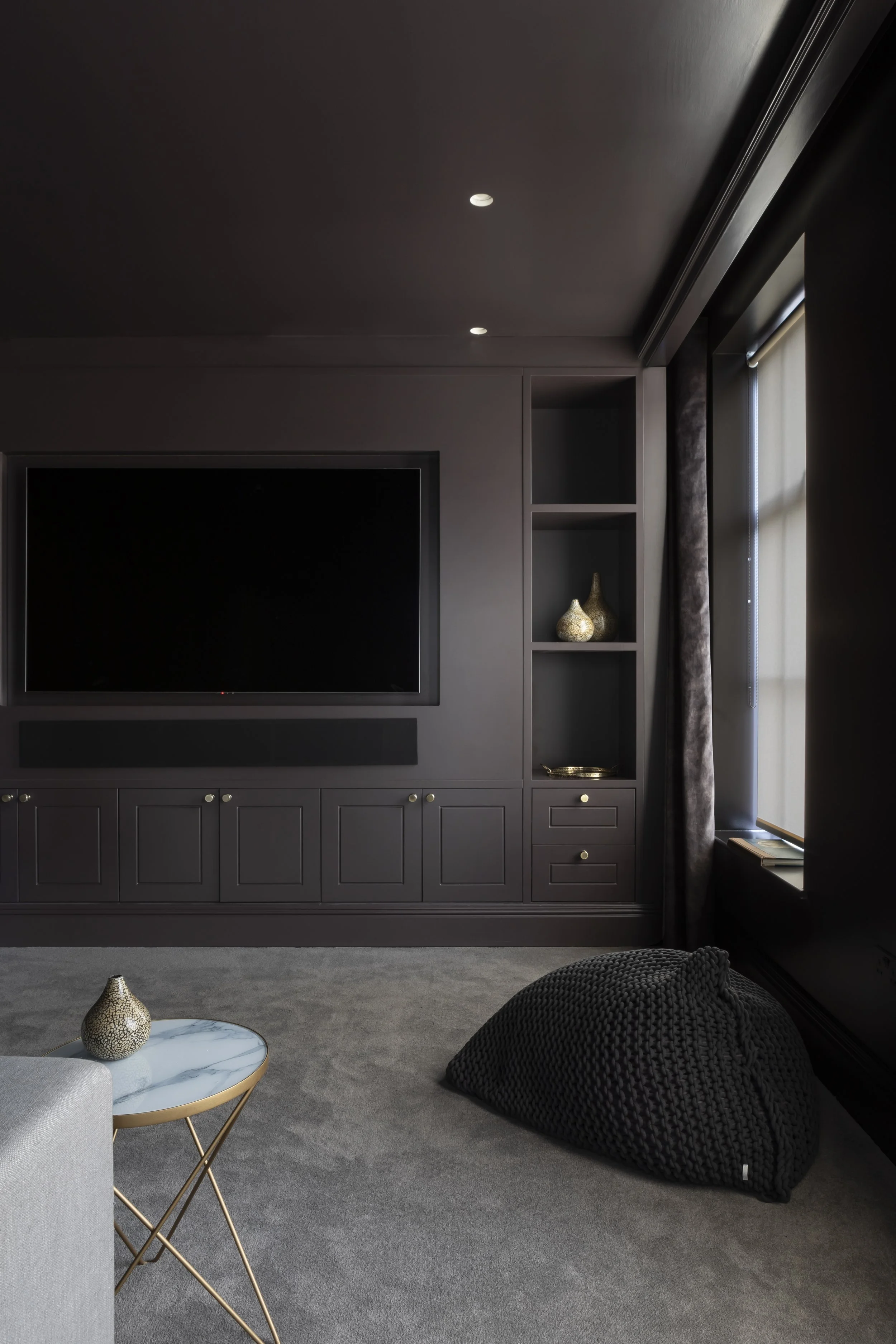 Modern living room with dark grey walls, large flat-screen TV, built-in shelves with decorative vases, window with curtains, grey carpet, black knitted beanbag, and side table with a decorative bowl.