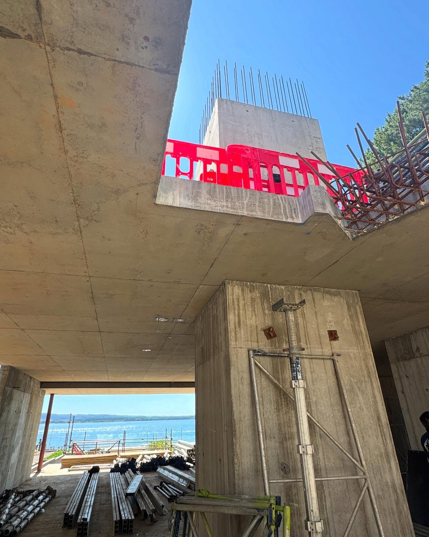 Construction site with concrete structure, steel reinforcement bars, and orange safety barrier, overlooking a body of water in the background.