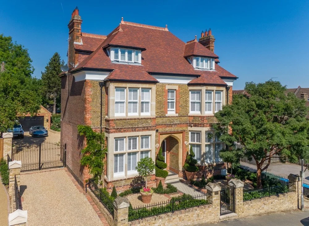 A large, multi-story brick house with a red-tiled roof, multiple chimneys, and white window shutters, surrounded by trees and a gated yard.
