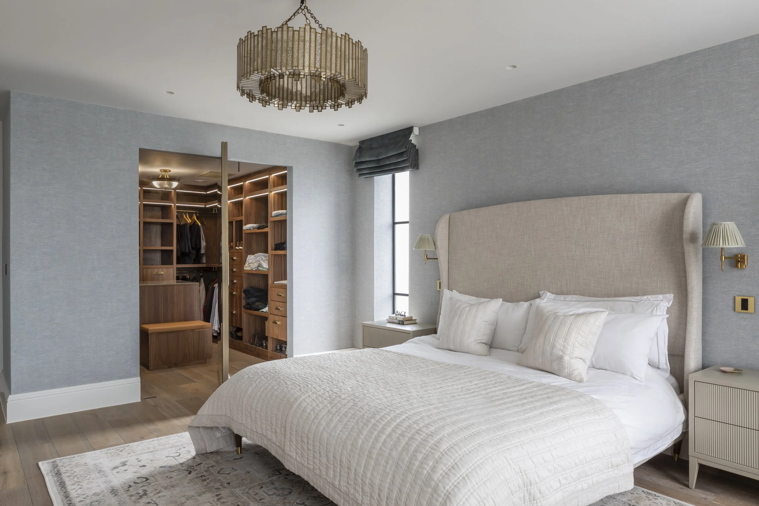 A modern bedroom with a large bed, beige upholstered headboard, white bedding, and two wall-mounted lamps. There is a closet with open wooden shelves and clothes, and a large window with dark valance drapes.