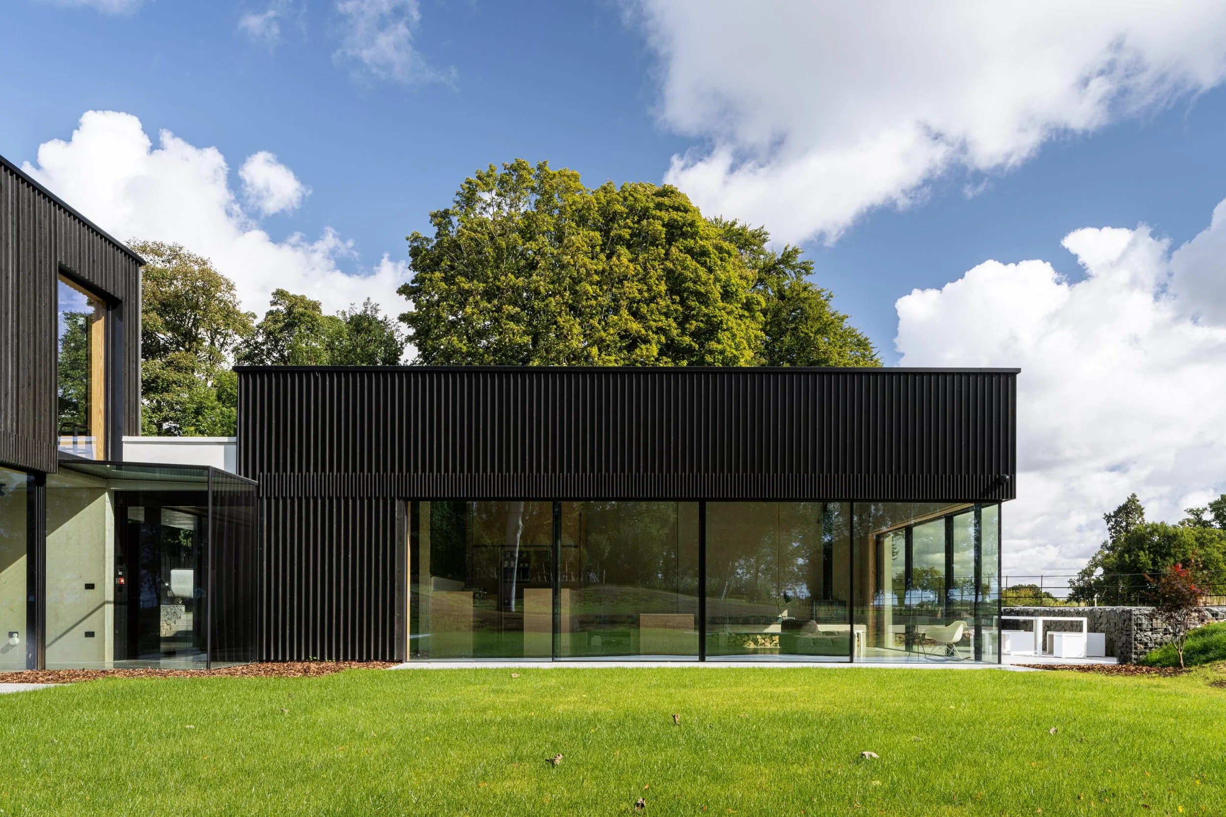 Modern house with black exterior walls and large glass windows, green lawn in foreground, tall trees and blue sky with clouds in background.