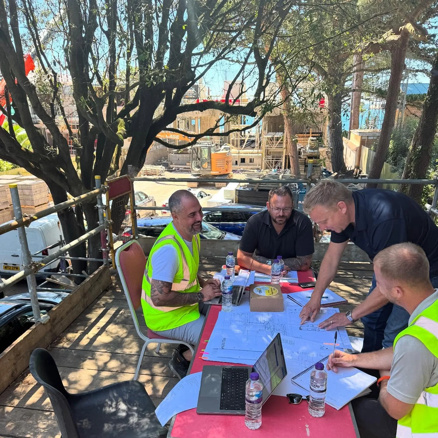 Group of construction workers and managers discussing plans at an outdoor site with trees, construction equipment, and partially built structures in the background.