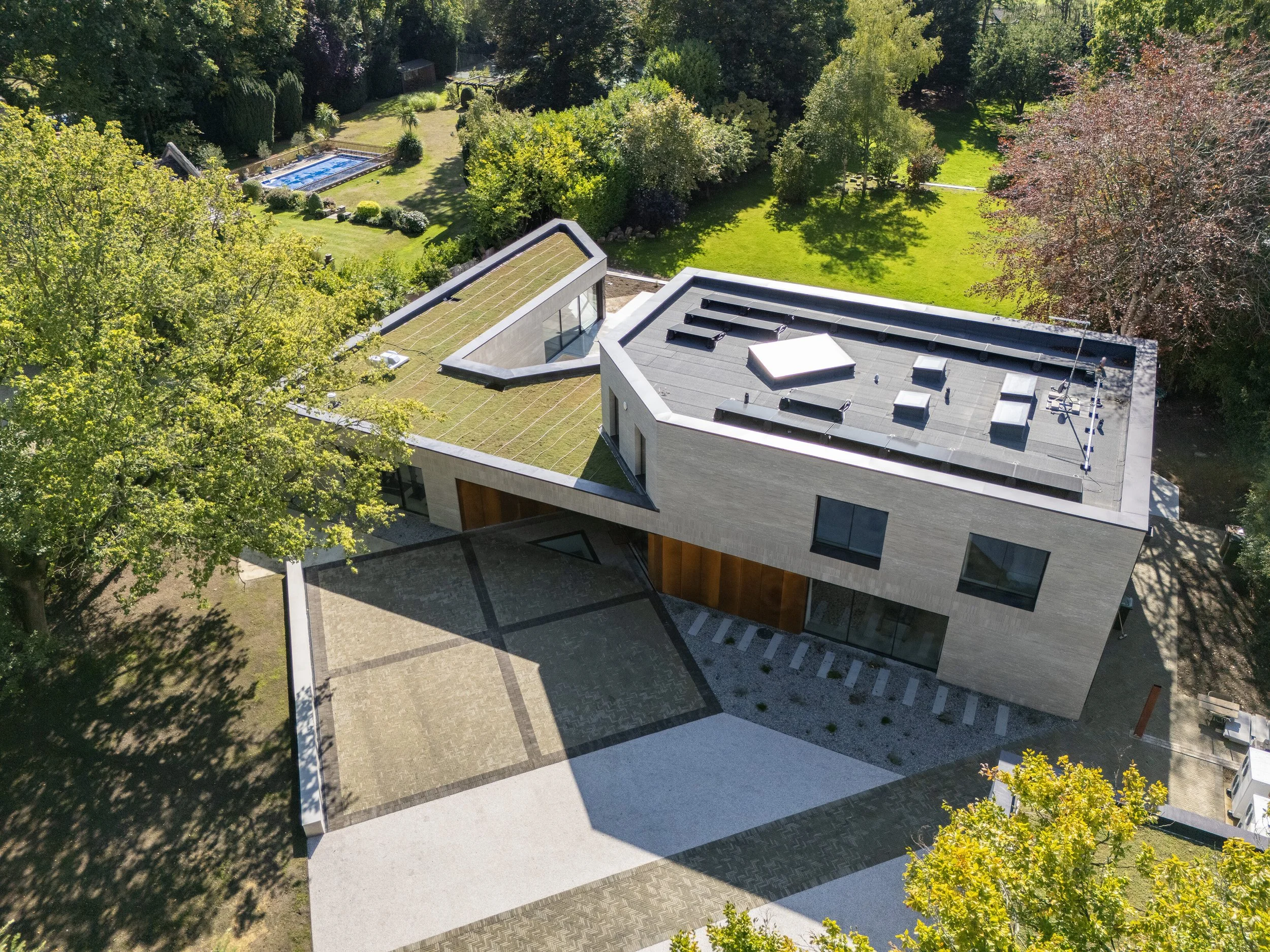Aerial view of a modern house with a green roof, large windows, surrounded by trees, and a backyard with a swimming pool and a patio.