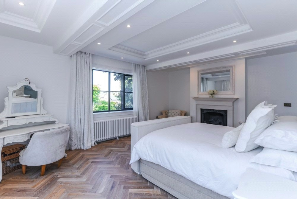 Bright bedroom with white walls, a large window with curtains, a white bed with multiple pillows, a fireplace with a mirror above, a vintage writing desk with a chair, a small armchair, and wooden herringbone flooring.