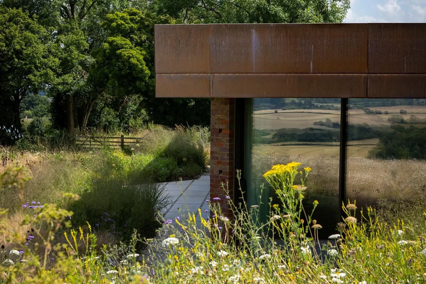 Modern house with large glass windows reflecting a green landscape and yellow flowers outside, surrounded by lush plants and trees.