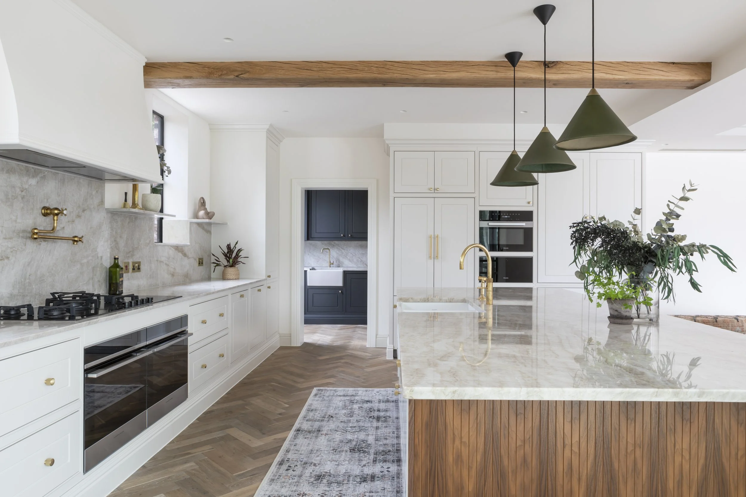 Modern kitchen with white cabinetry, marble countertops, a large kitchen island with a marble top, gold fixtures, hanging green pendant lights, wooden accents, and potted plants.