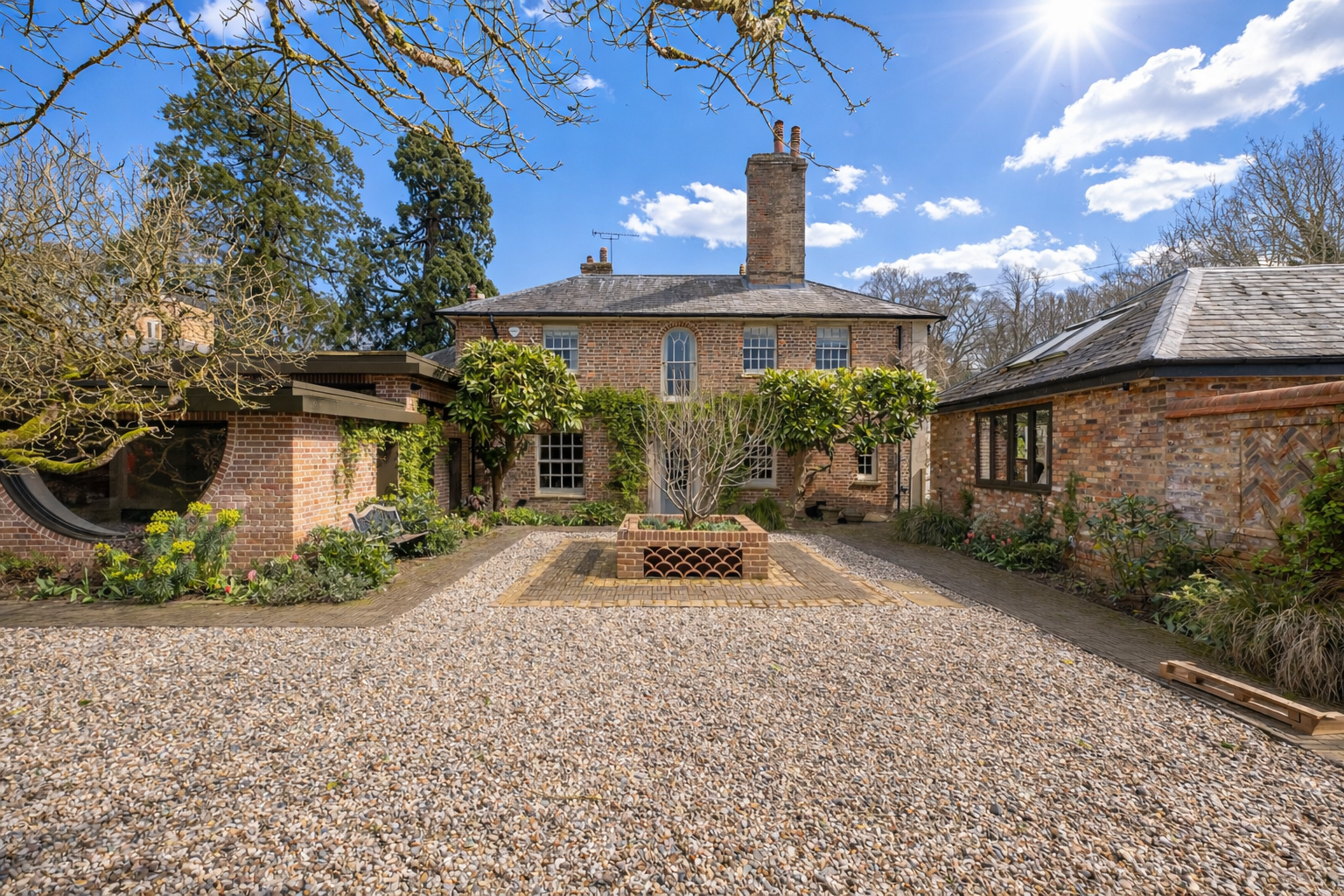 A brick house with a courtyard featuring a tree in a raised brick planter, surrounded by gravel and brick pathways, and a blue sky with clouds overhead.
