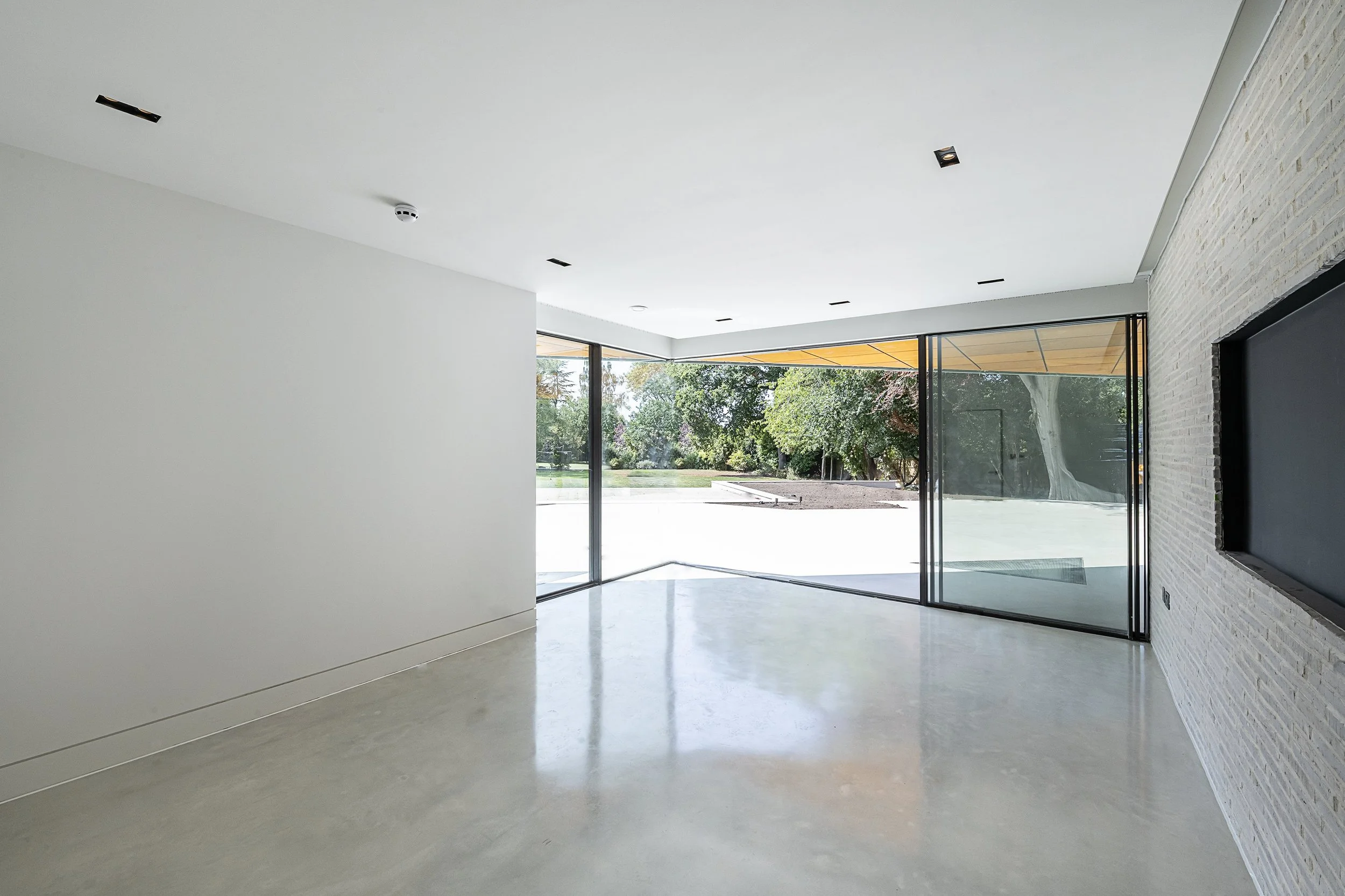 Empty modern living room with white walls, polished concrete floor, and large glass sliding doors opening to outdoor garden.