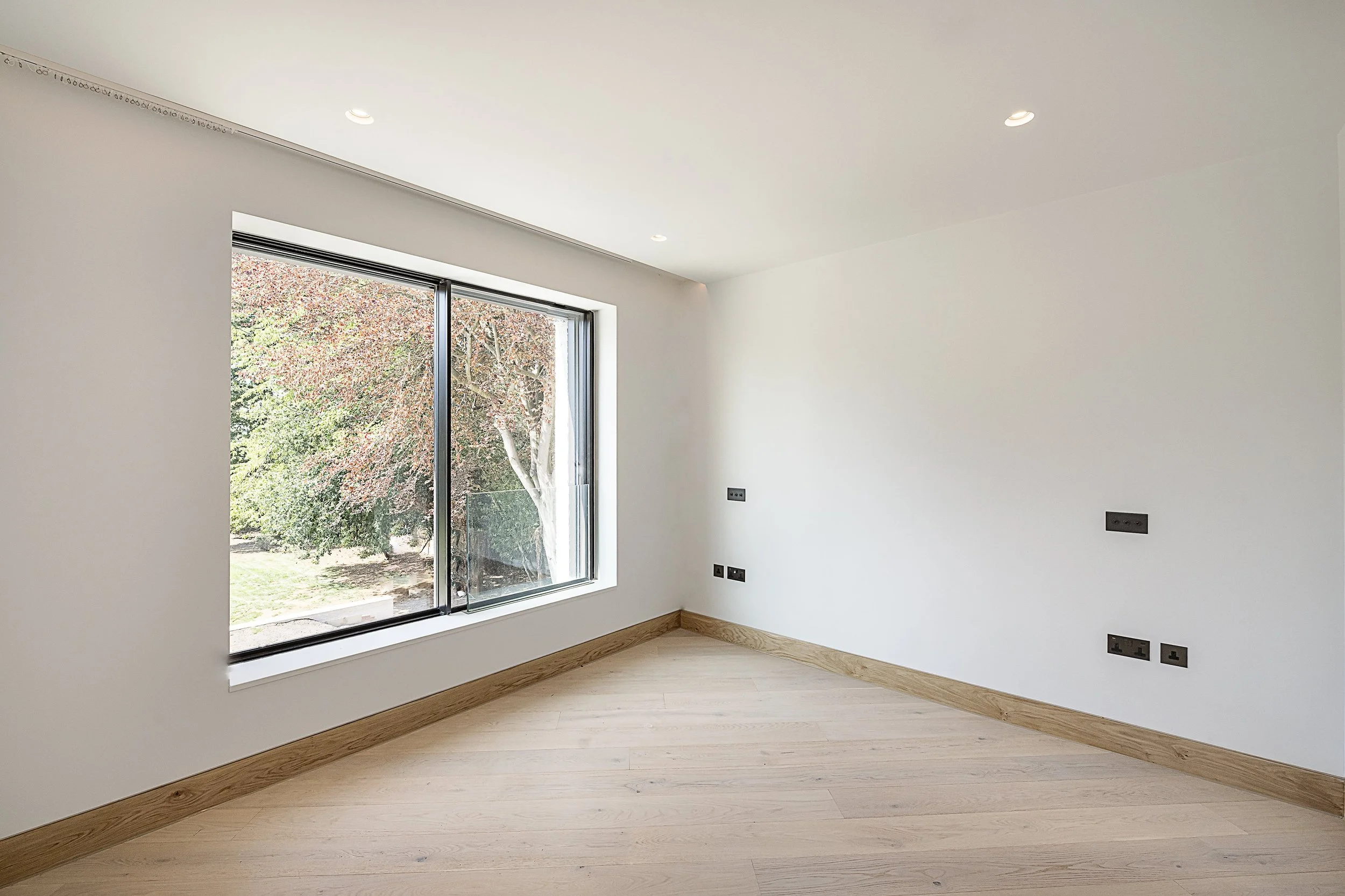 Empty room with white walls, hardwood floor, large window showing trees outside, ceiling with recessed lighting, and multiple electrical outlets.