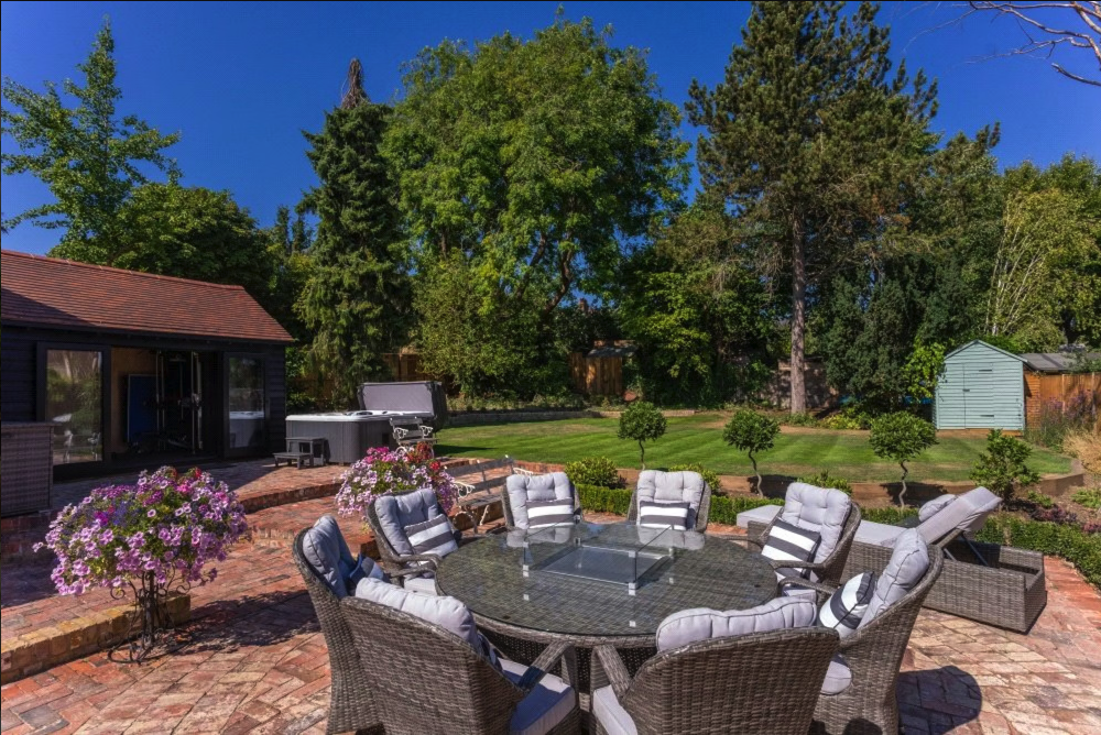 Outdoor patio with a round wicker table and cushioned chairs, a hot tub, blooming flowers, lush green lawn, and trees under a blue sky.