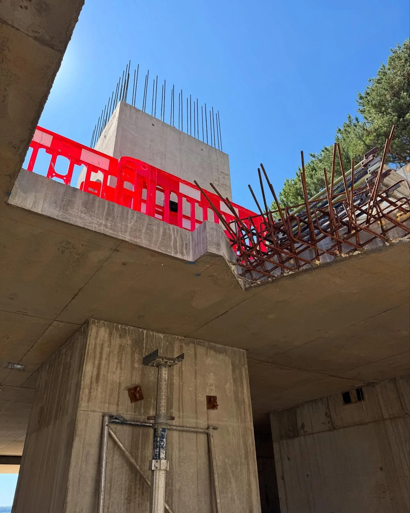 View of a construction site showing concrete structure, steel reinforcement bars, and safety barriers, with trees and a blue sky in the background.