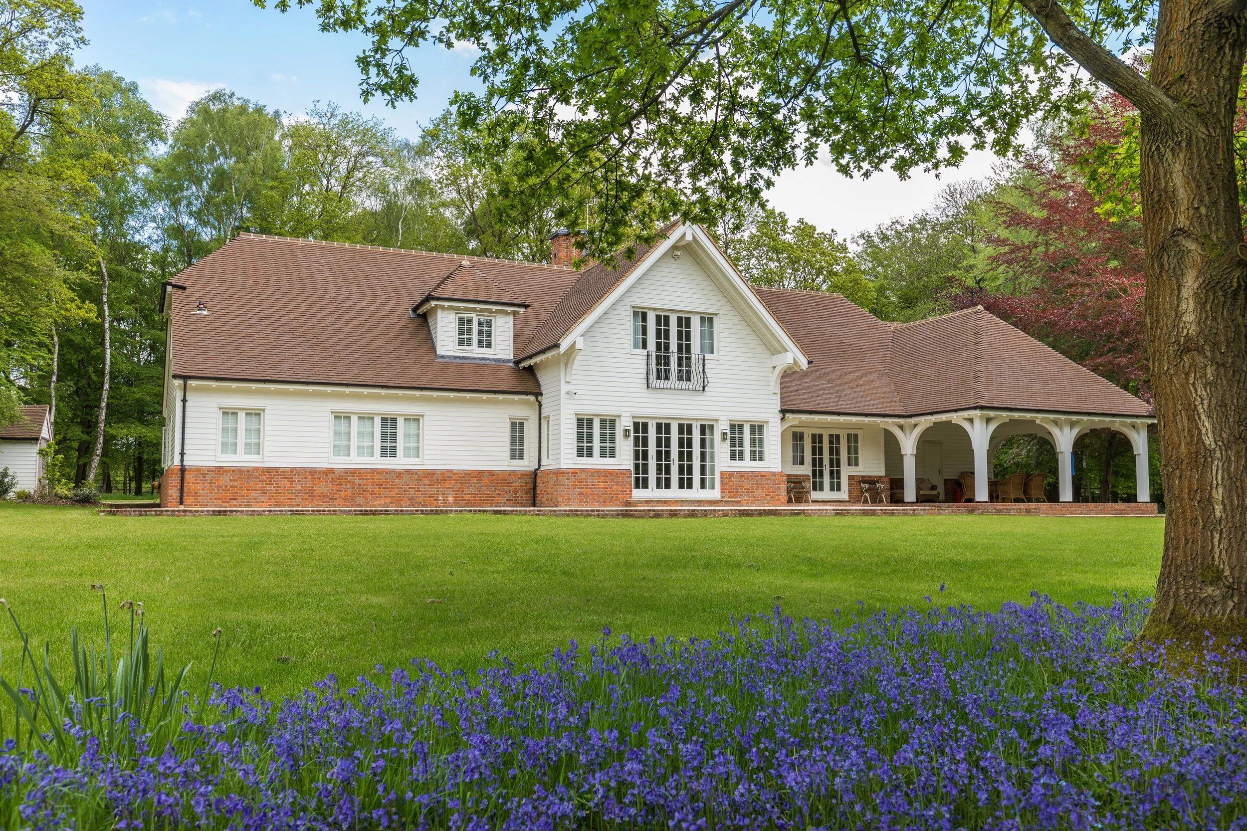 A large white house with a brown shingled roof, brick foundation, and a covered porch, surrounded by a green lawn and trees, with purple flowers in the foreground.