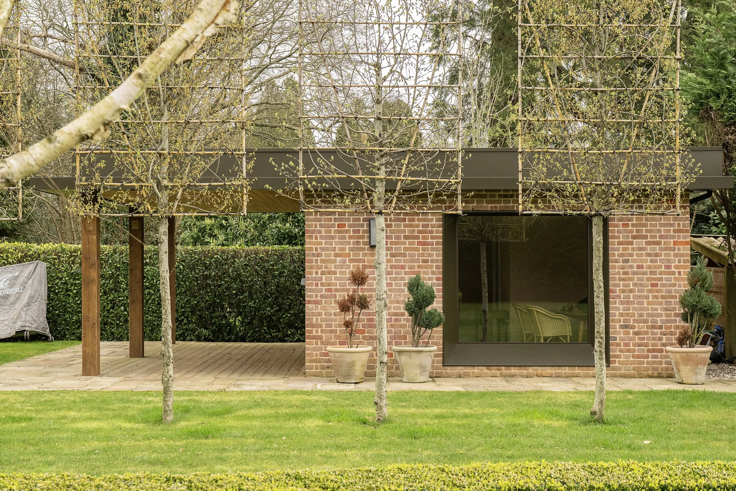 A modern brick house with a large window and a deck, surrounded by trees and potted plants, with a green lawn in front.