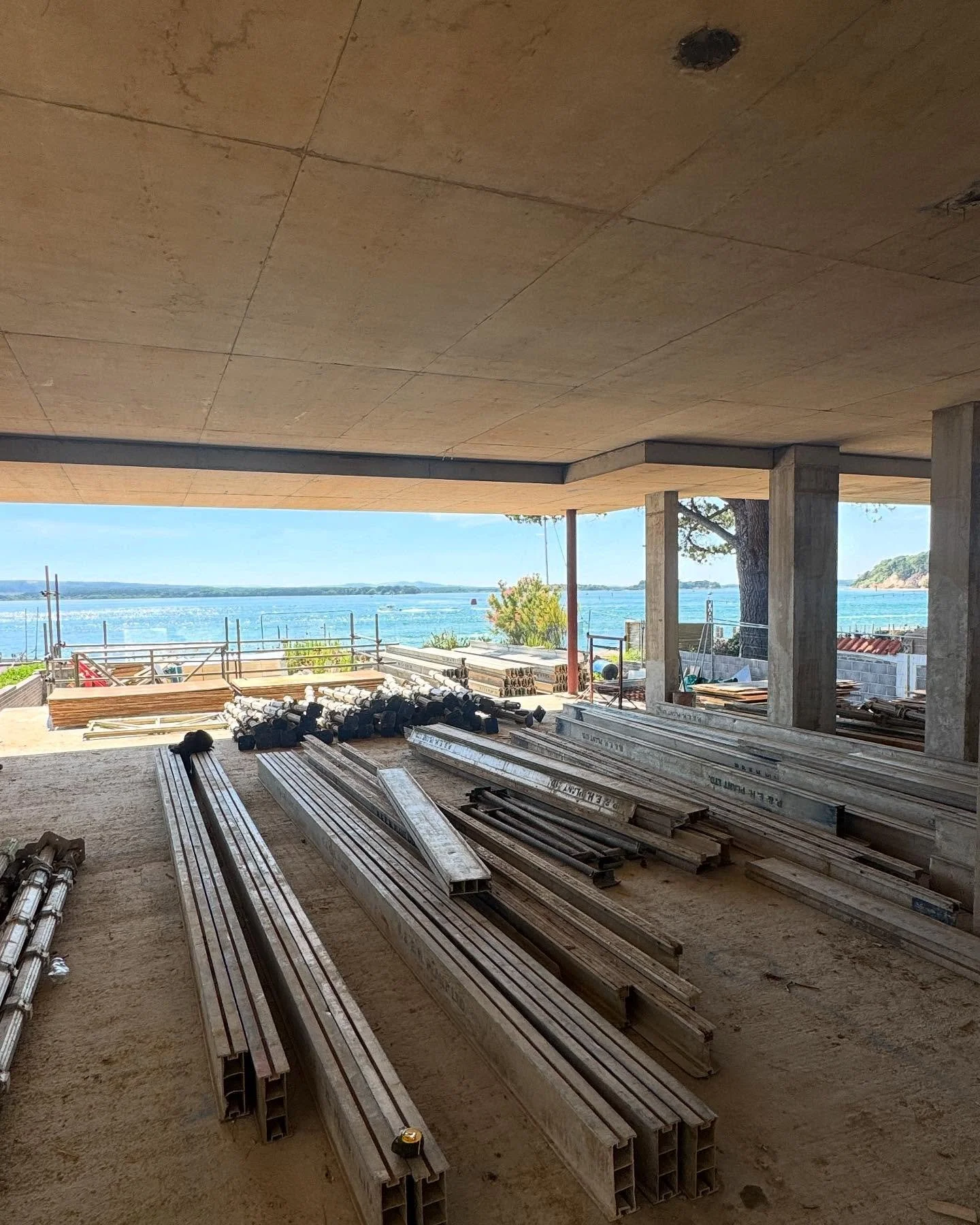 Construction site with metal beams and scaffolding overlooking a body of water and distant islands under a partially constructed building.