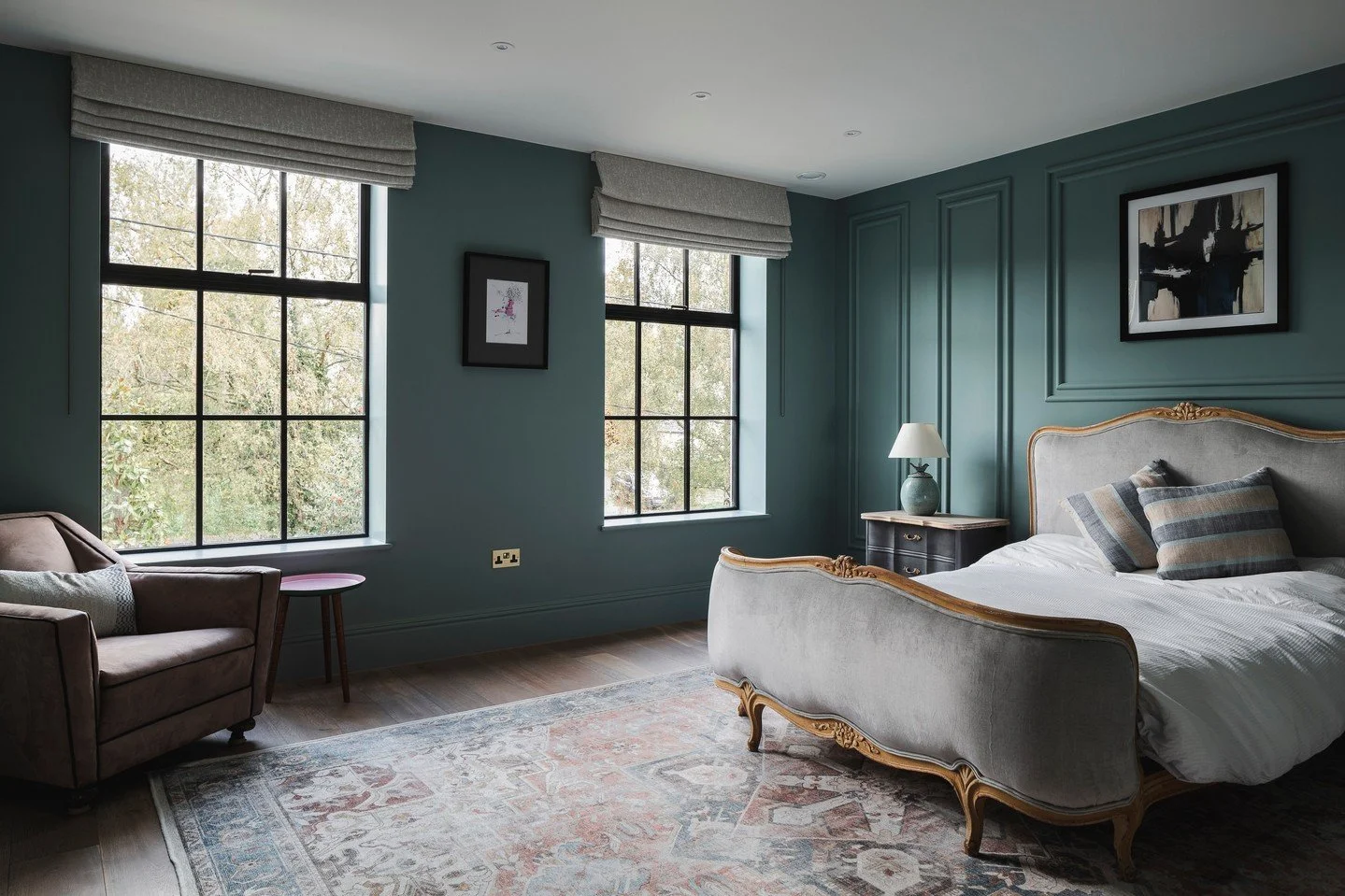 A bedroom with dark teal walls, tall windows with beige curtains, a vintage cream-colored bed with wooden trim, a black nightstand with a white lamp, a patterned area rug, and an armchair near the window.