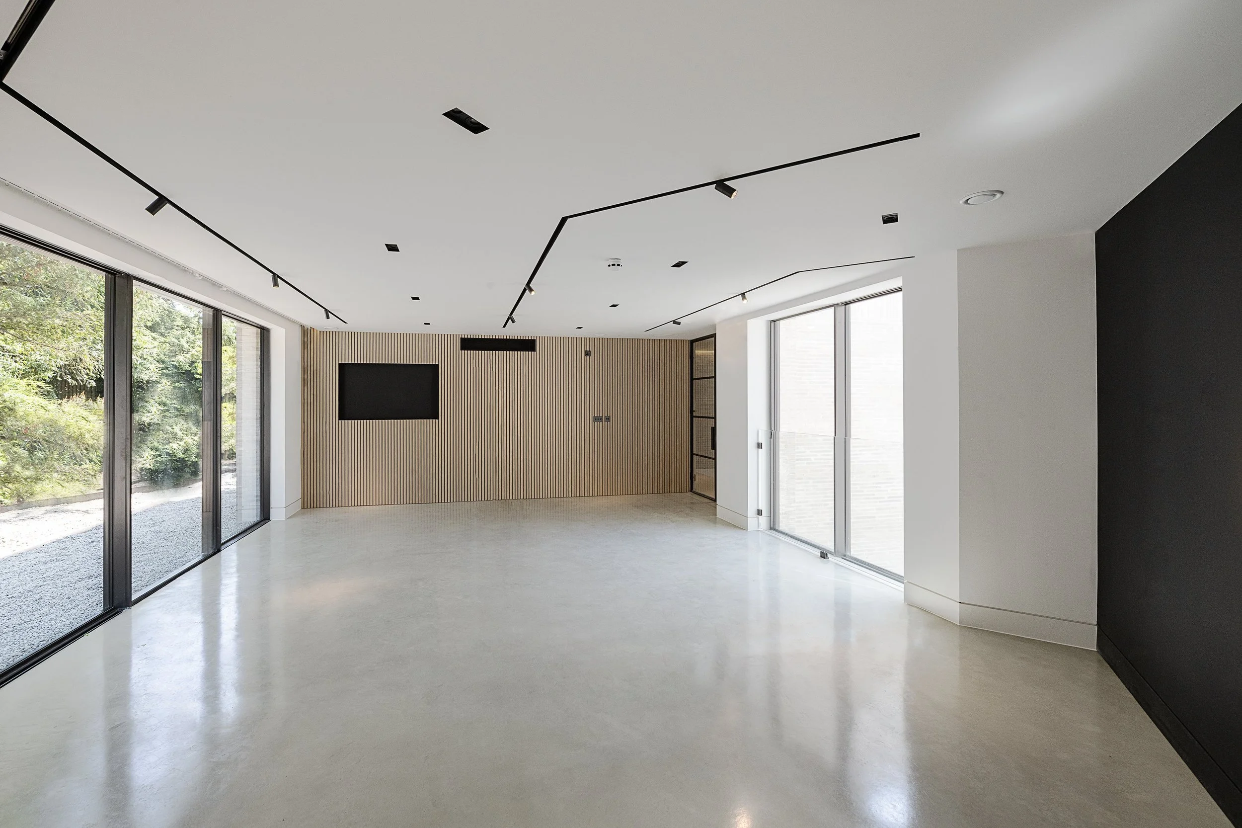 Empty modern room with large windows, light-colored floor, black ceiling accents, and a wooden wall with a mounted television.