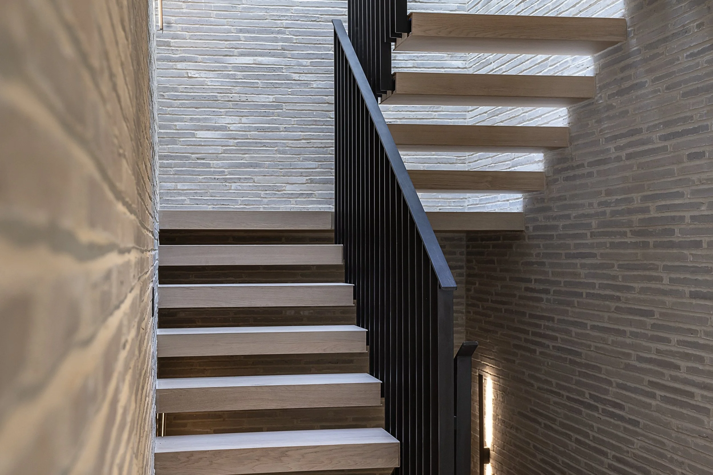 Modern staircase with wooden steps and black railing in a room with brick walls.