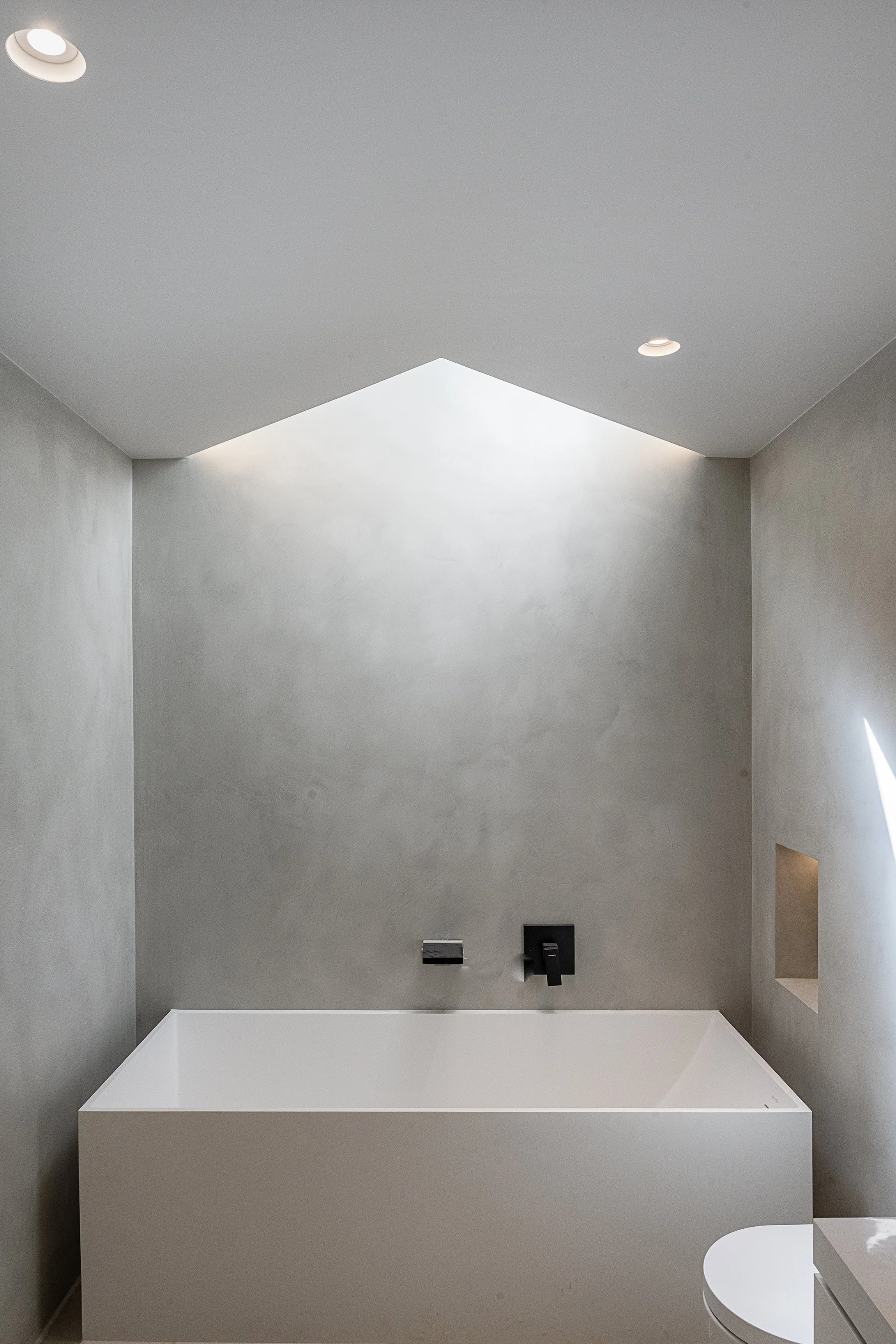Modern minimalist bathroom with a built-in bathtub, gray textured walls, a skylight ceiling, and a small square window on the right wall.
