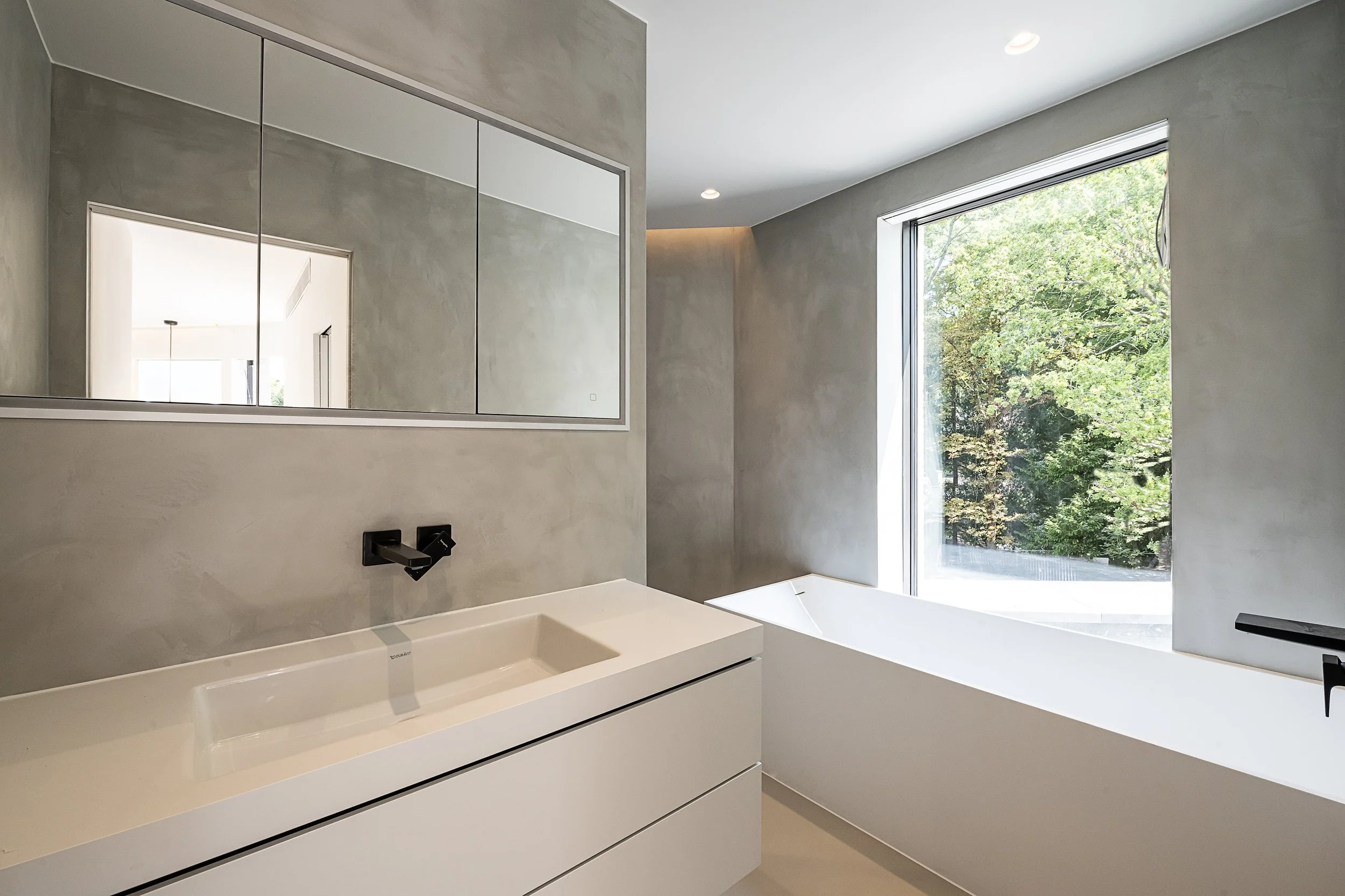 Modern bathroom with a large window, white bathtub, minimalist sink, and a wall mirror cabinet.