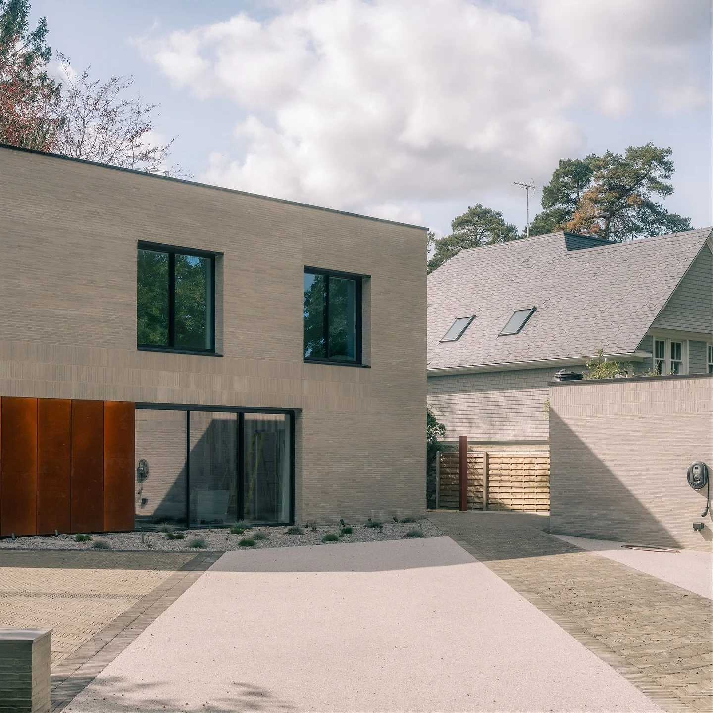 Modern beige brick house with large glass sliding doors on ground level, two black-framed windows above, and a gravel and brick driveway. Neighboring traditional house with sloped roof and solar panels. Fence with wooden gate in background. Clear sky